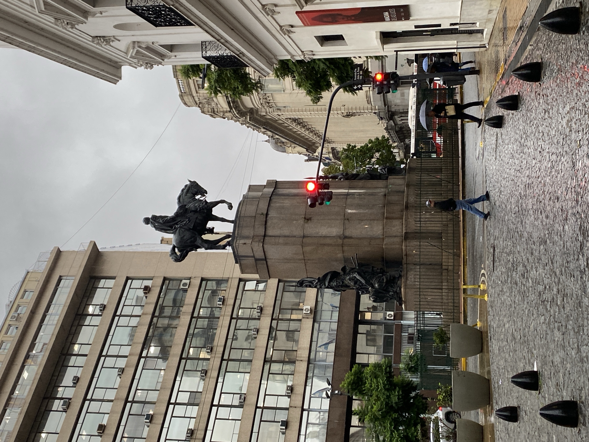 The image depicts a street scene in Buenos Aires, Argentina, during a rainy day. The street is wet and reflects light. There is a large equestrian statue atop a stone monument, which is a prominent landmark in the area. A modern building with many windows is visible behind the monument. The traffic lights are red. People are walking on the sidewalk, some carrying umbrellas. Black bollards are present along the edge of the sidewalk. The architecture includes both classical and modern styles. There are trees and planters lining the street.