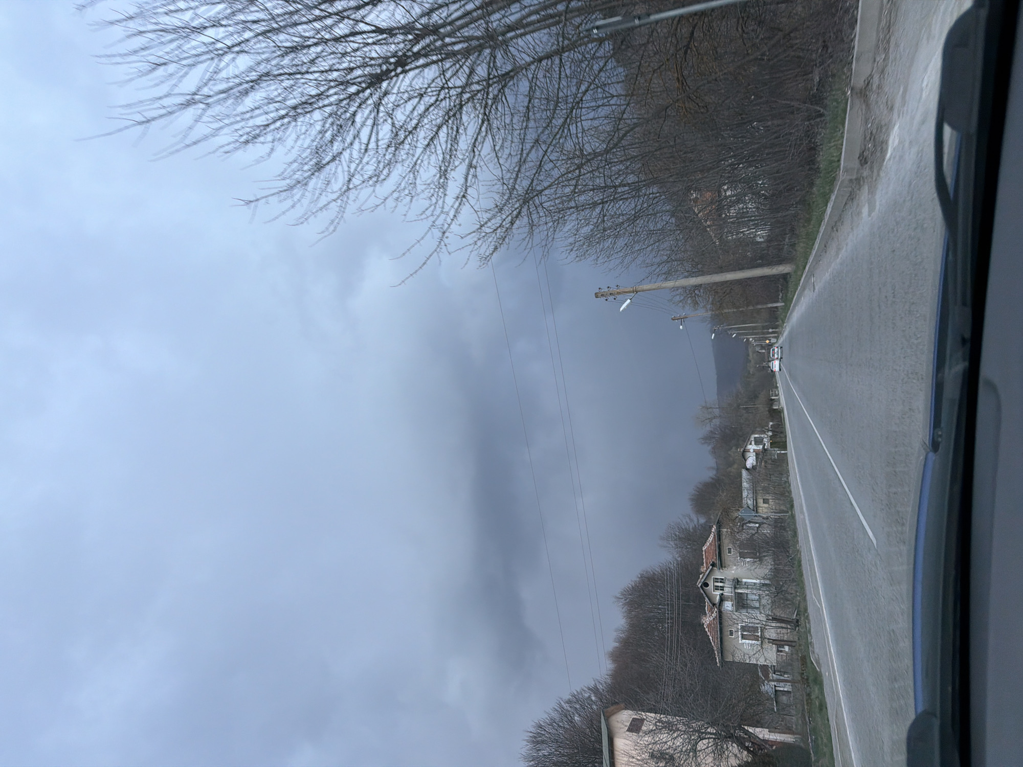 The image depicts a road in the village of Gintsi, Bulgaria, under a heavily overcast sky. The road is wet, likely from recent rain. A white dividing line runs down the center of the road. On the right side of the road are houses, most appearing somewhat dilapidated. Trees, bare of leaves, line the roadside. Utility poles and wires are visible along the road. The view is likely taken from inside a vehicle, as part of a dashboard is visible on the right side of the frame. No people are visible. A couple of cars appear far off down the road, but they lack detail.