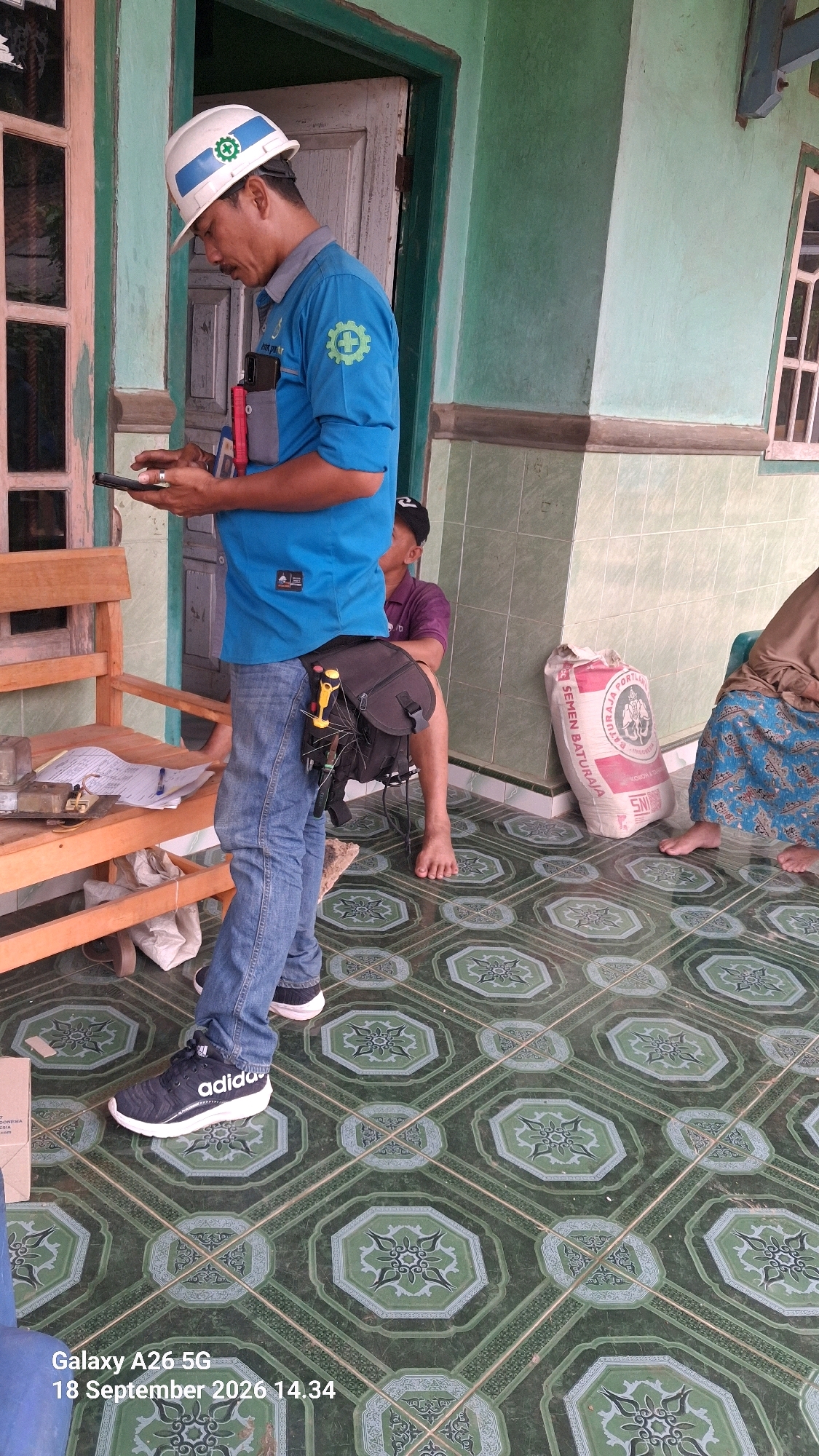 An Indonesian man wearing a blue uniform, a white hard hat with a green logo, and blue jeans is standing on a patterned tiled floor. He is holding a dark smartphone in his left hand and looking down at it. He has a tool belt around his waist with various tools attached, including a yellow-handled screwdriver. To his right, another person wearing a purple shirt and a black cap is sitting on a stool, partially visible. Further to the right, a sack of cement with Indonesian text and a green logo is leaning against a wall. In the background, there is a building with green and white walls and a wooden door. The scene appears to be outdoors on a porch or veranda. The timestamp "Galaxy A26 5G 18 September 2026 14.34" is superimposed on the bottom left of the image.