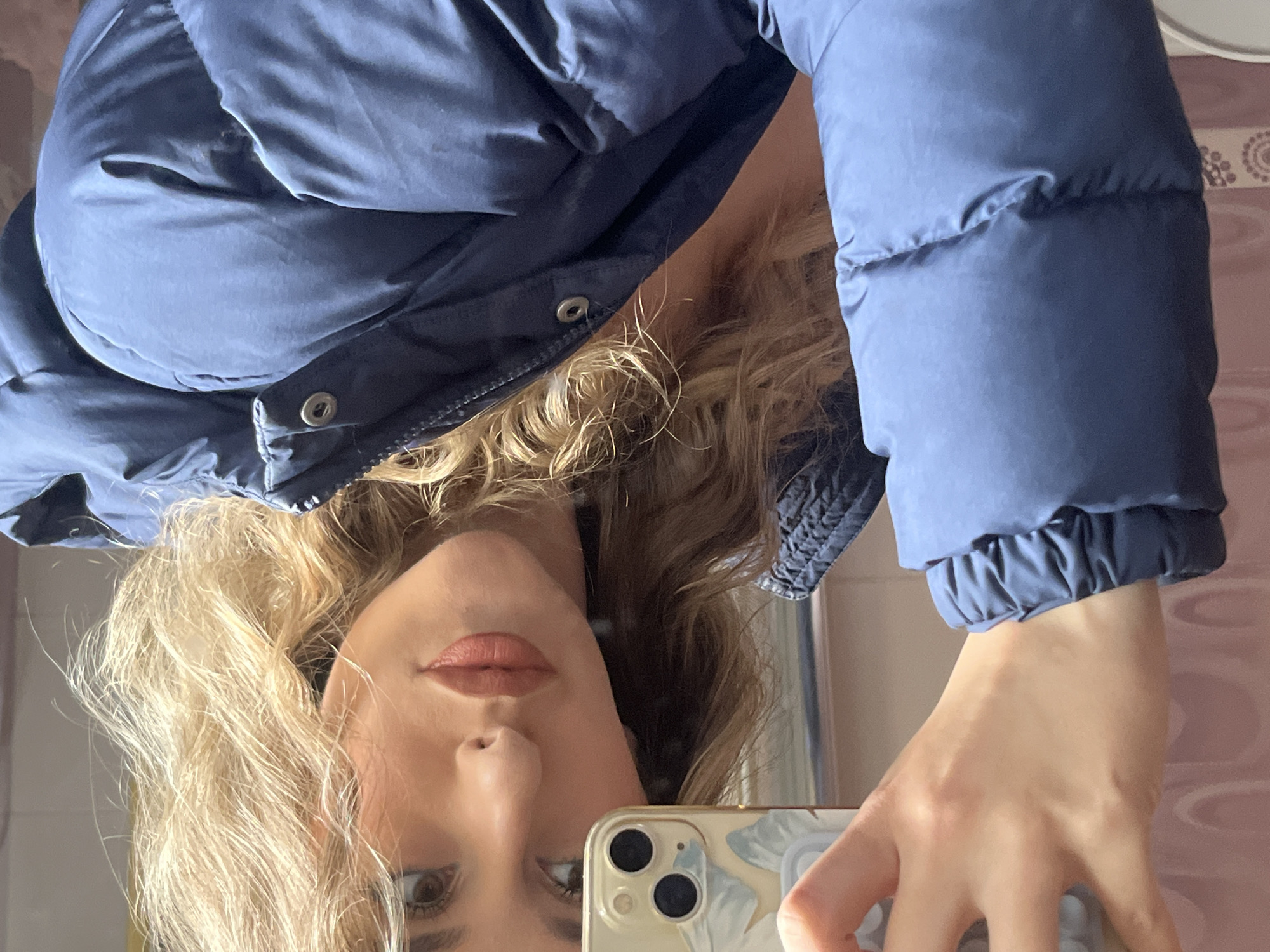 A woman is taking a selfie in a mirrored surface. She is wearing a blue puffer jacket. The reflection of a bathroom wall with a light pink patterned design is visible in the mirror. The location is indeterminate as there are no distinctive visual elements to place the scene in Sofia, Bulgaria.