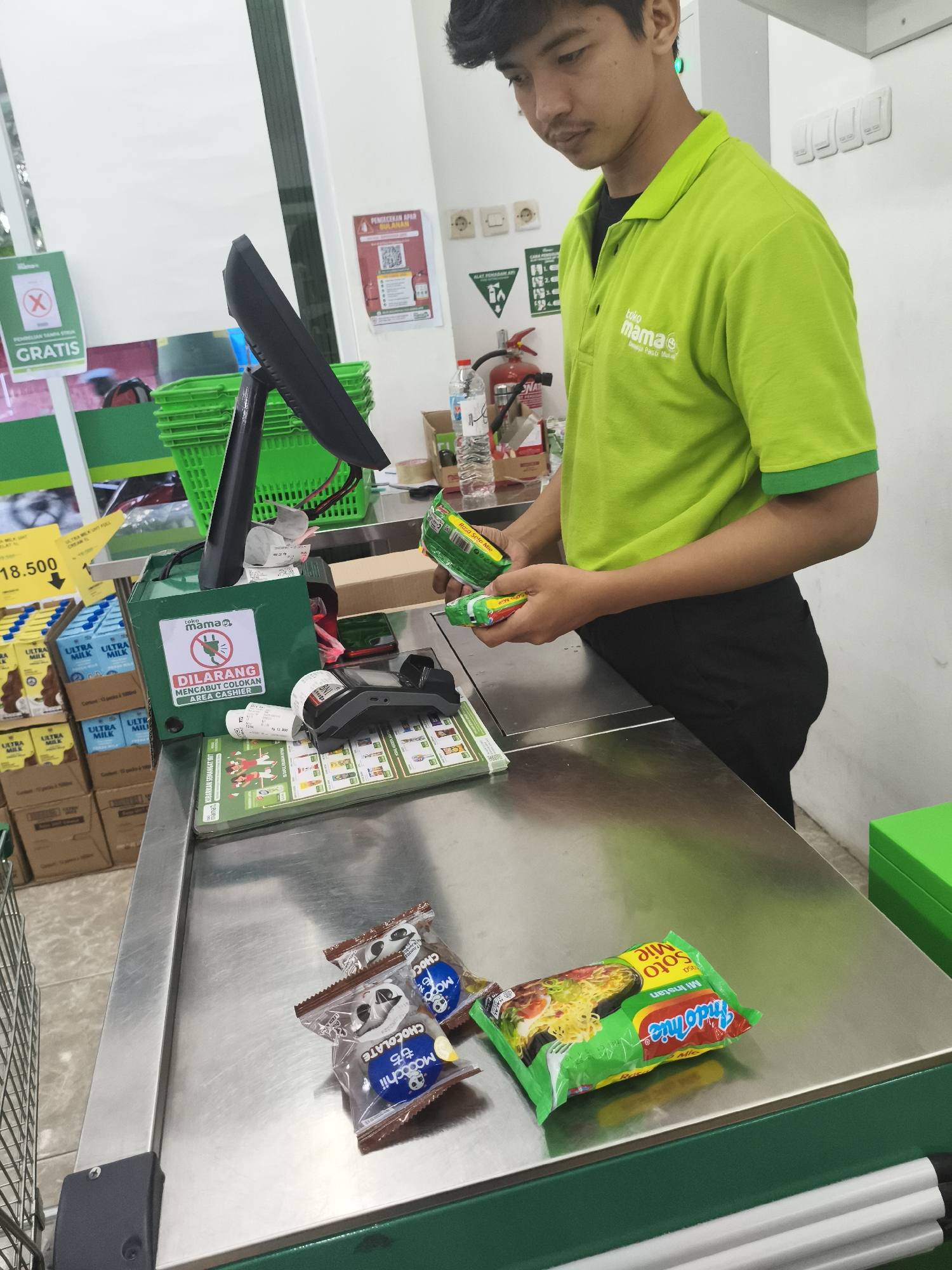 A young man in a bright green polo shirt with the "Toko Mama" logo is standing behind a checkout counter in a supermarket. He is holding two items, one green and one yellow, and appears to be in the process of scanning them. On the counter are several other items, including two small bags of chocolate snacks and a package of instant noodles. A cash register and payment terminal are also visible. The background shows shelves stocked with products, a shopping cart, and signs indicating promotions and store policies. The overall scene depicts a typical grocery store checkout in Depok, Indonesia.