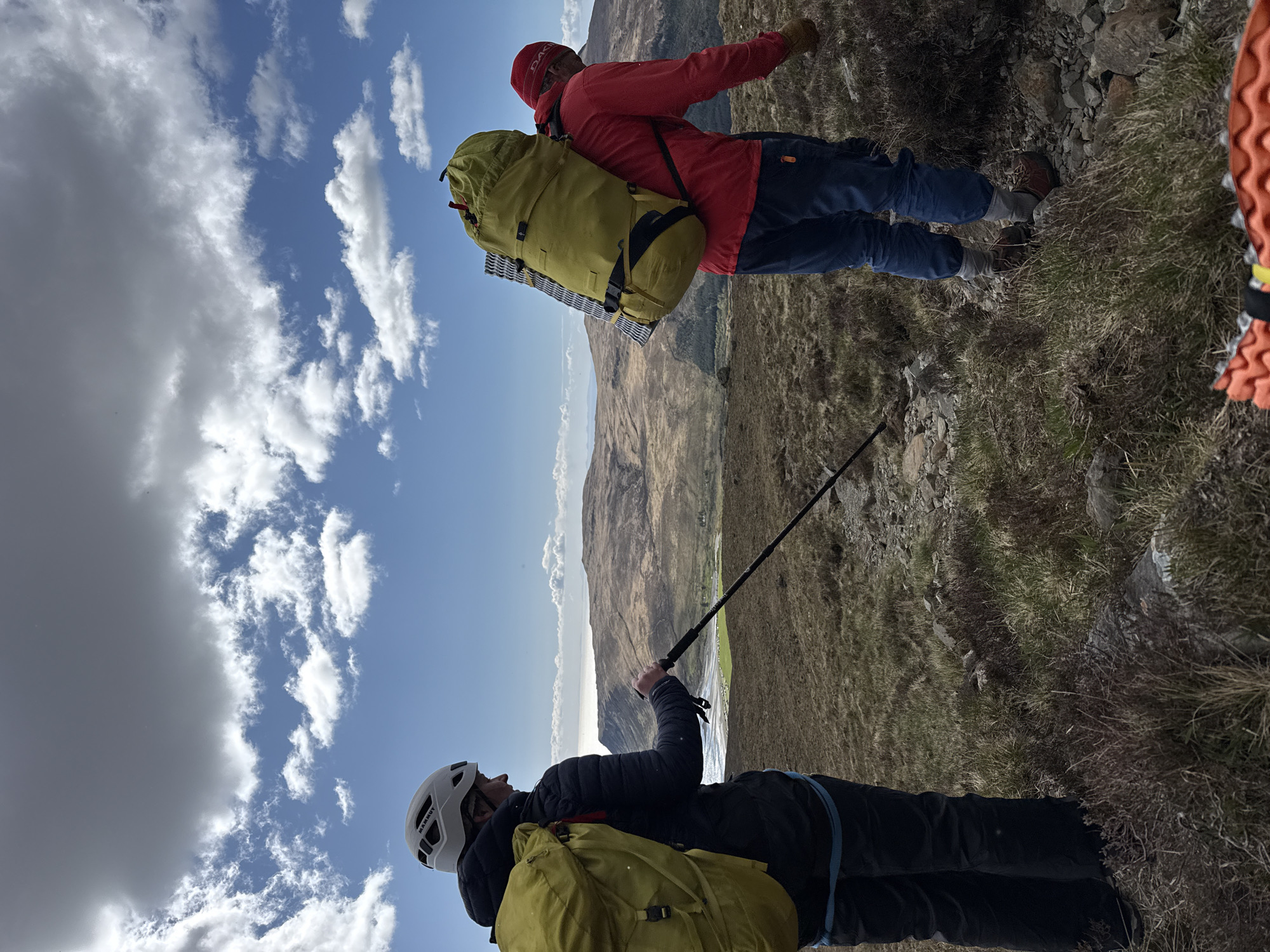 Two individuals are depicted in a mountainous outdoor environment. The lower individual, wearing a white helmet, a dark jacket, and carrying a yellow backpack, holds a walking stick. The upper individual is dressed in a red jacket and blue pants, also carrying a yellow backpack with a rolled mat attached. The background features a landscape of mountains under a sky with scattered clouds. The location is Unknown, Unknown.