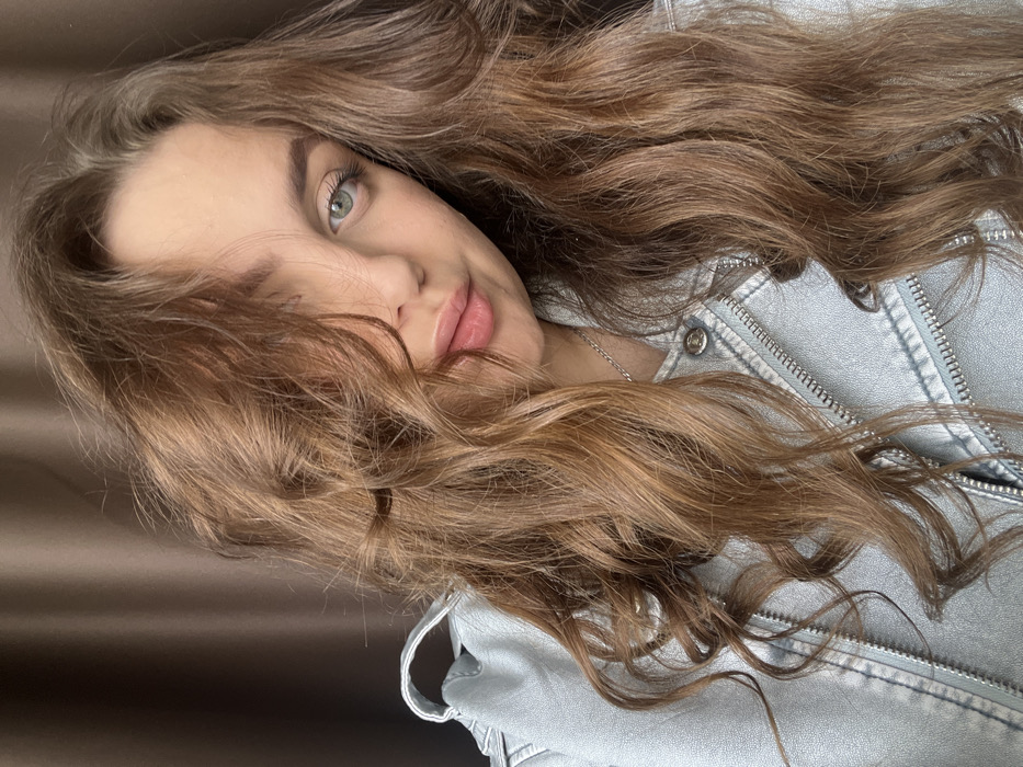 A single female subject is depicted in a close-up portrait, focusing on her head and upper torso. Her long, wavy, light brown hair is prominent, partially obscuring the right side of her face (viewer's left). Her visible eye is light-colored, appearing greenish-blue. She has full lips with a pink tint and distinct eyebrows. The subject is wearing a light blue denim jacket featuring visible white stitching and silver zippers. A thin silver chain necklace is visible around her neck. She maintains direct eye contact with the viewer, exhibiting a slight, closed-mouth smile. The background is a plain, dark brown or taupe color, consistent with an indoor setting, possibly a curtain or wall. No specific identifying features of the city of Chaikovsky, Russia, are discernible within the image frame.