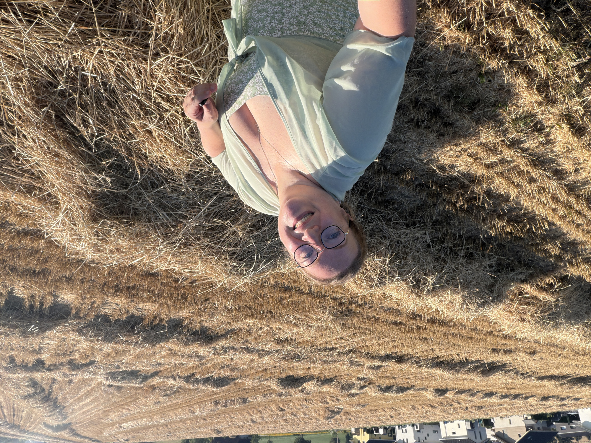 In Ufhusen, Switzerland, a woman stands in a harvested wheat field, with rows of stubble and straw laid out before her. She is wearing a green floral dress and a light green sheer kimono-style cardigan. She has on glasses and a necklace, and her hair is short and blonde. Behind her, a row of houses and trees can be seen in the distance. The lighting suggests it is daytime, with the sun casting shadows across the field.