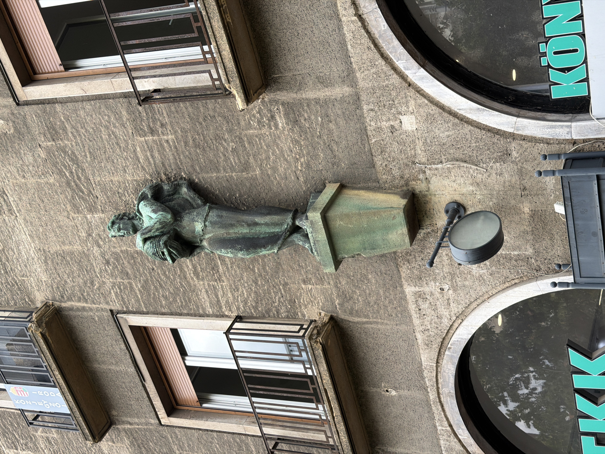 This is a low-angle, medium shot of a bronze sculpture of a human figure on the exterior of a building in Debrecen, Hungary. The sculpture is green with patina and depicts a person holding a round object, possibly a beehive, in their arms. The figure stands on a weathered green pedestal that is mounted on the building's stone facade.
Above the sculpture, there are two windows with balconies. The left window has a rolled-down brown shutter, and the right window shows an open shutter revealing a white interior. Both balconies have black metal railings and stone ledges, with the left ledge showing signs of wear and damage. A banner with Hungarian colors and text hangs from the left balcony.
Below the sculpture and to the left, part of a large, arched window or storefront is visible, with the green neon letters "TKK" illuminated. To the right of the arched window, the green neon letters "KÖNY" are visible on another storefront. A circular, dark-colored sign hangs from an articulated arm on the building's facade between the sculpture and the arched windows. The building's facade is constructed of rough-hewn stone with visible horizontal striations. The overall impression is of an urban architectural setting.