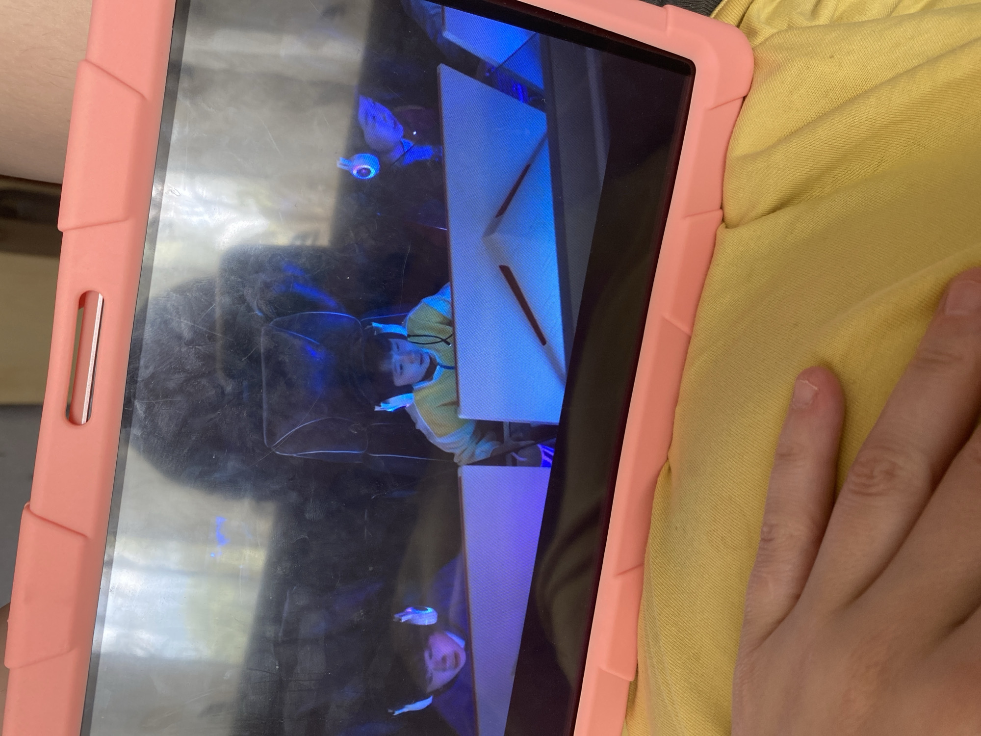 A close-up shot shows a tablet in a pink case, resting on a yellow fabric surface. The tablet screen displays a scene with two young children wearing headphones and playing what appears to be a video game. The children are positioned behind futuristic-looking gaming setups with blue and purple lighting. In the foreground, a person's hand, likely the viewer's, is visible, resting on the yellow fabric. The image is taken indoors, and the background on the tablet screen suggests an artificial, possibly gaming-related environment. The overall scene is casual and domestic, with no specific city or location details identifiable.