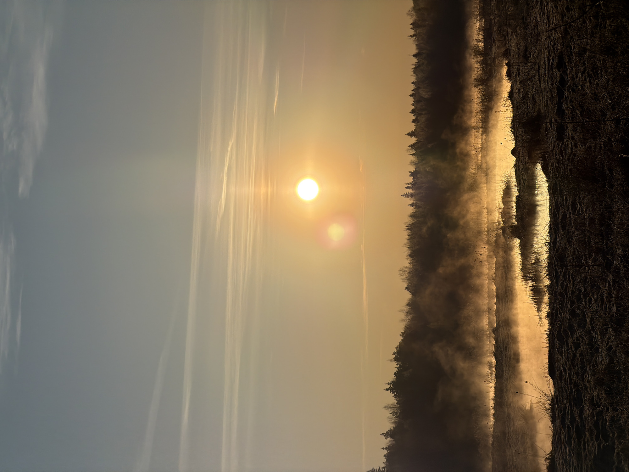 The image depicts a landscape at sunrise or sunset, with a body of water bisecting the scene. On the right side of the water, there is a line of trees bordering the shore. The water is covered in a layer of fog or mist, creating a hazy atmosphere. The sun is visible in the center of the image, casting a golden light across the sky and water. The left side of the image shows a clear sky with some thin clouds. The location is identified as being in Izhevsk, Russia.