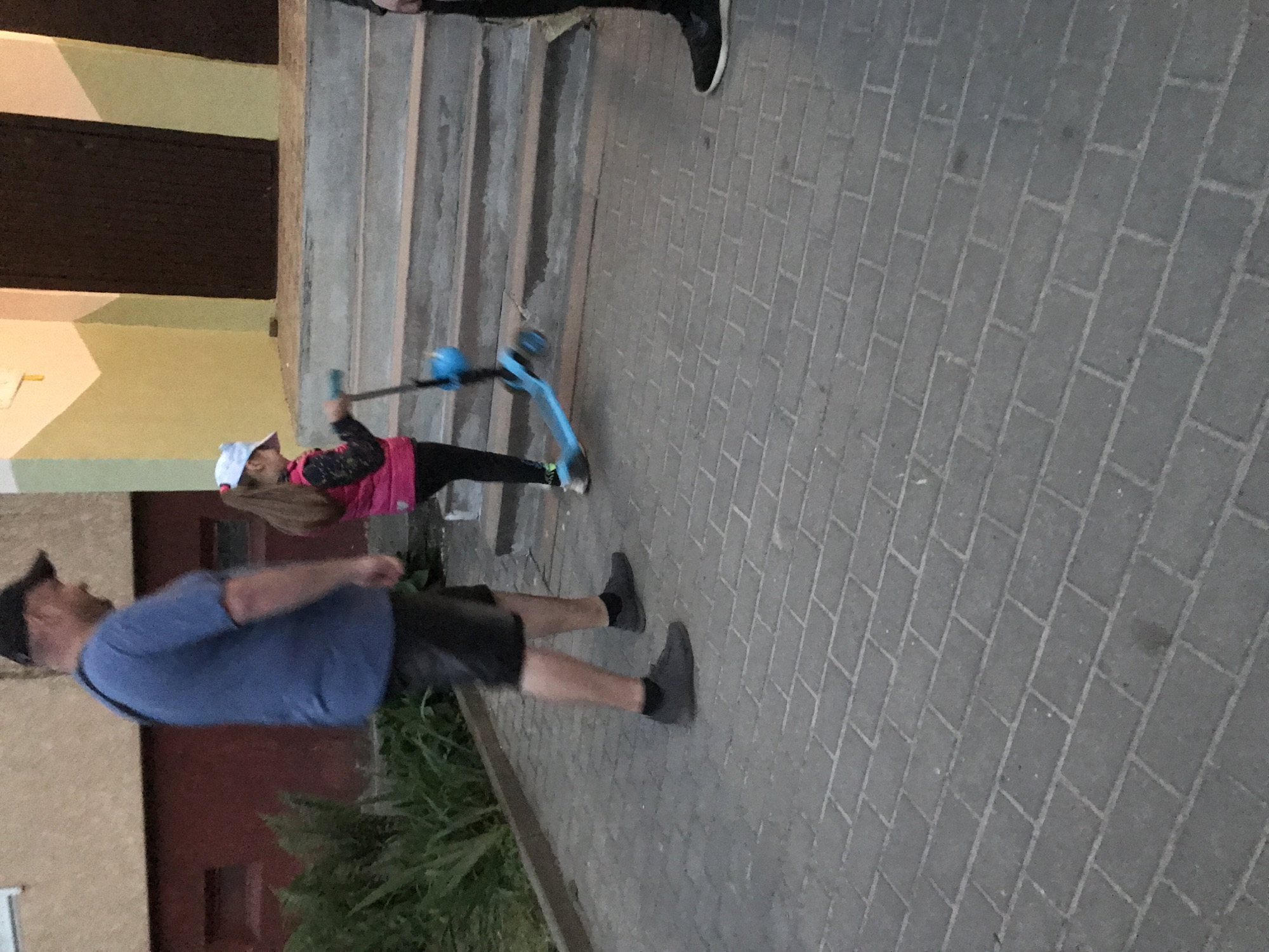 This outdoor image, taken in Ternopil, Ukraine, depicts a casual scene during what appears to be late afternoon or early evening, characterized by soft, ambient lighting. The ground is uniformly paved with grey rectangular interlocking stones.
In the foreground on the left, an adult male stands, facing slightly to his right. He is dressed in a blue short-sleeved t-shirt, dark shorts, a dark baseball cap, and dark sneakers paired with dark socks. His arms are bent, and his hands are not clearly visible.
Towards the center-right of the frame, a young girl is present, holding onto a bright blue kick scooter. She has long brown hair and is wearing a pink long-sleeved top with a dark pattern, black leggings, a white baseball cap, and light-colored shoes. She seems to be interacting with her scooter, possibly preparing to ride or resting.
In the upper right background, only the lower legs and feet of another individual, wearing black sneakers with white soles, are partially visible on a set of concrete or stone stairs that ascend towards the upper right.
The background on the left features a building with a light-colored facade, possibly beige or pale yellow, interspersed with dark rectangular elements that could be windows or doors. Some dark green foliage or bushes are visible in the lower left corner, bordering the paved area. The scene conveys a calm, everyday moment with no strong emotions discernible. There is no visible text in the image.