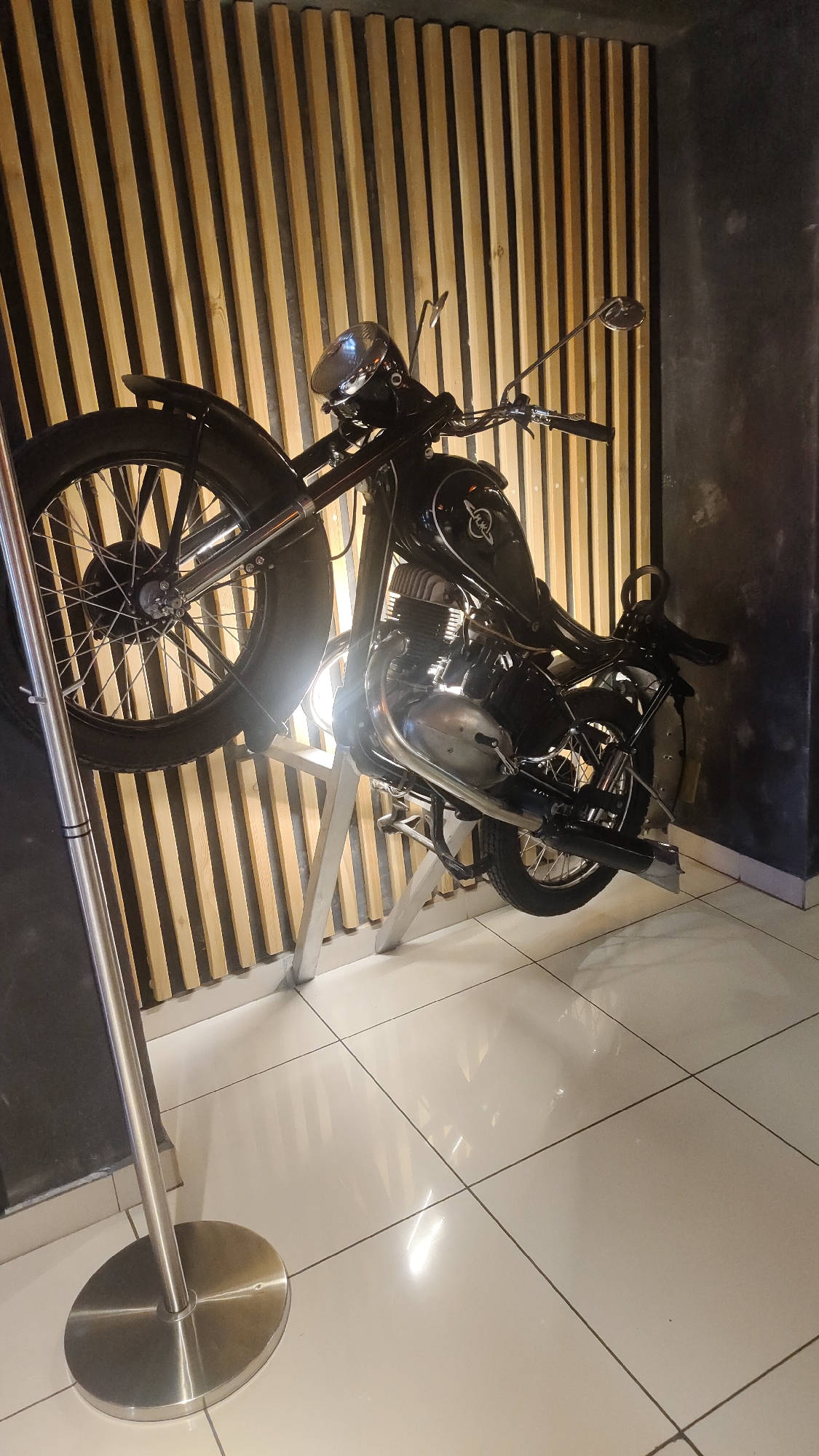 The image shows a black vintage motorcycle displayed in an indoor setting. The motorcycle is mounted on a silver-colored stand, slightly tilted to the right. It features a large front wheel with spokes, a headlight, a fuel tank with lettering, and a solo seat. The background consists of a wall with vertical wooden slats on the left and a dark-painted wall on the right. The floor is tiled in light squares with a reflective surface. A silver pole and circular base stand are positioned to the left of the motorcycle. Based on search results, this is in the restaurant "Kochevnik", located in Krasnoyarsk, Russia.