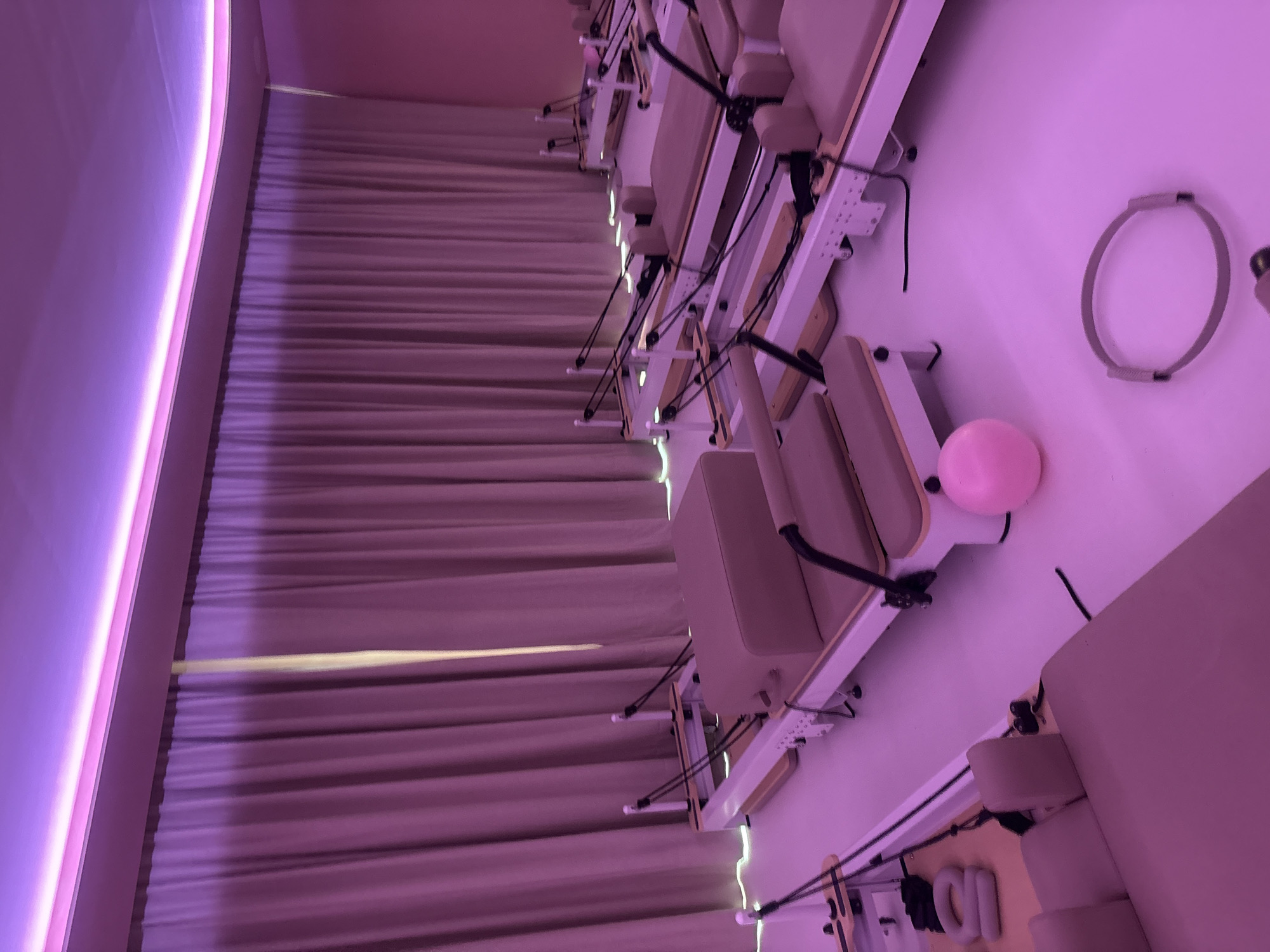 A pilates studio in Mexico City, Mexico, features a row of reformers aligned against a wall draped with floor-to-ceiling curtains. The studio is illuminated with pink and purple light. A Pilates ring and a small exercise ball are positioned on one of the reformers.