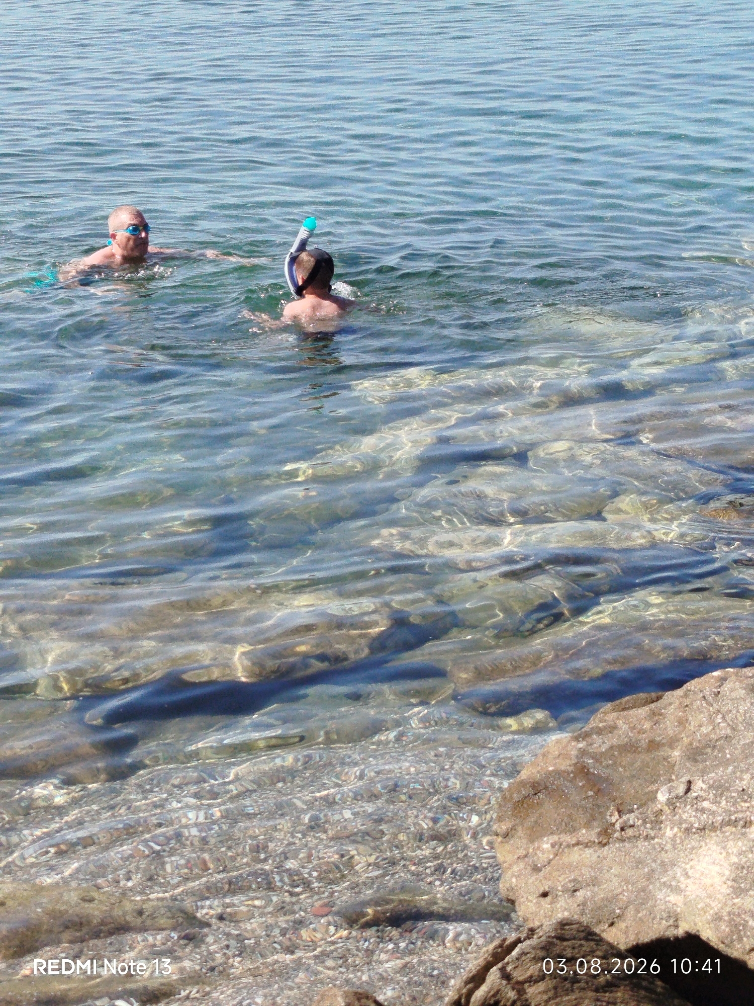 In Sithonia Municipal Unit, Greece, two individuals are engaged in snorkeling in clear, shallow water. The person on the left, an adult male with a shaved head and blue goggles, is facing towards the right. To his right, another individual, possibly a child or younger person, is wearing a snorkel mask and appears to be swimming. The water is exceptionally clear, revealing the seabed which is covered in small stones and pebbles. The sunlight refracts through the water, creating shimmering patterns on the surface. In the foreground, a large, rough-textured rock juts out of the water on the right side of the frame. The overall scene depicts a serene moment of recreational activity in a coastal environment.