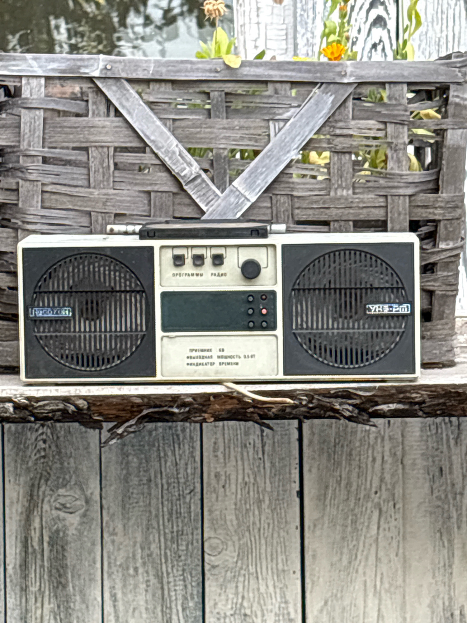 The image displays a vintage portable radio positioned on a rustic wooden surface, possibly a shelf or fence. The radio is off-white and beige, with black speaker grilles on either side. The left speaker grille has a label that appears to be in Cyrillic script. The center panel of the radio features control buttons, a circular dial, and a small dark green display. Text in Cyrillic is visible below the display, indicating "PRIEMNIK KB" (receiver KB), "VYKHODNAYA MOSHCHNOST 0.5 VT" (output power 0.5 W), and "INDIKATOR VREMENI" (time indicator). The radio is placed in front of a woven wooden structure that resembles a basket or screen, possibly with plants or flowers visible behind it. The background shows dappled light and greenery. The wooden surface in the foreground is made of vertical planks, showing grain and texture. The overall scene has an outdoor, possibly aged aesthetic. The provided image is not from Bratsk, Russia.
