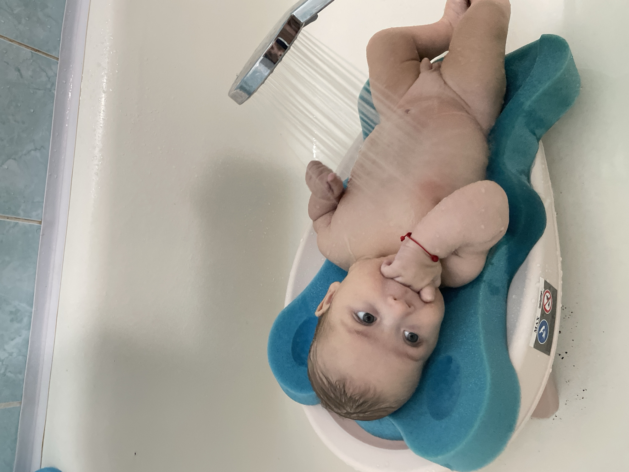 The media file depicts a baby being bathed in a bathtub. The baby is lying on a blue bath support, which is placed in a white bathtub. A shower head is visible at the top left of the image, with water spraying down onto the baby. The baby has light brown hair and is looking at the camera with a curious expression. The baby is wearing a red bracelet on its left wrist. The background shows a portion of a tiled wall in a light blue color. There are some water droplets on the surfaces. A sticker is visible on the side of the bath support, with some text and symbols. One symbol appears to be a warning sign. Another symbol looks like a checkmark within a circle. The word "MAX" is also visible on the sticker. The scene appears to be indoors, in a bathroom. The lighting suggests it could be daytime.