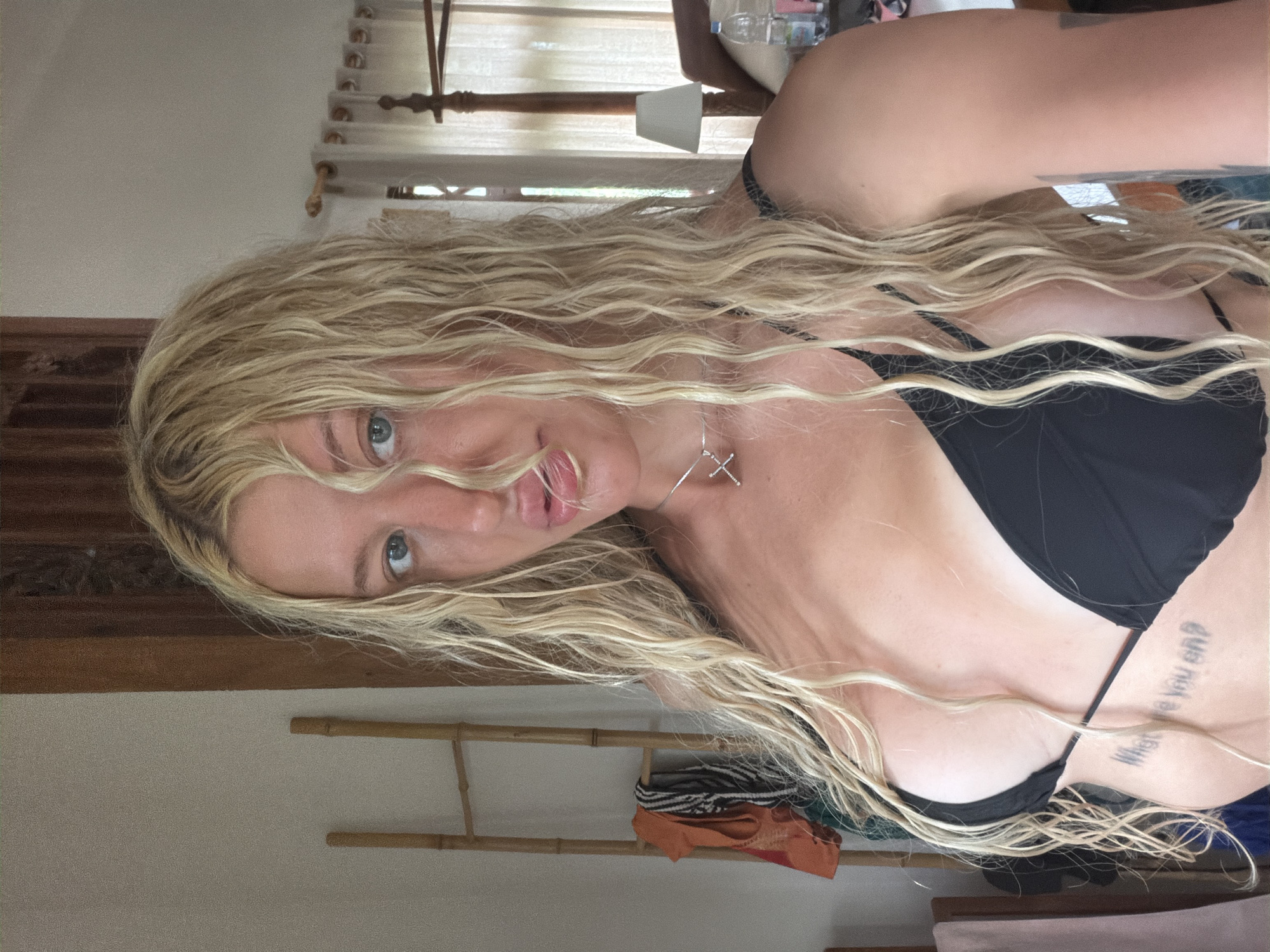 A young woman with long, blonde, wavy hair stands in what appears to be a bedroom. She is wearing a black bikini top and a necklace with a silver pendant. Her hair is partially covering her face. In the background, there is a wooden ladder with clothes hanging on it, sheer curtains, a bed with a wooden headboard, and a bedside table with a lamp and a bottle of water. The room appears to be a hotel or guesthouse room in North Kuta, Indonesia.