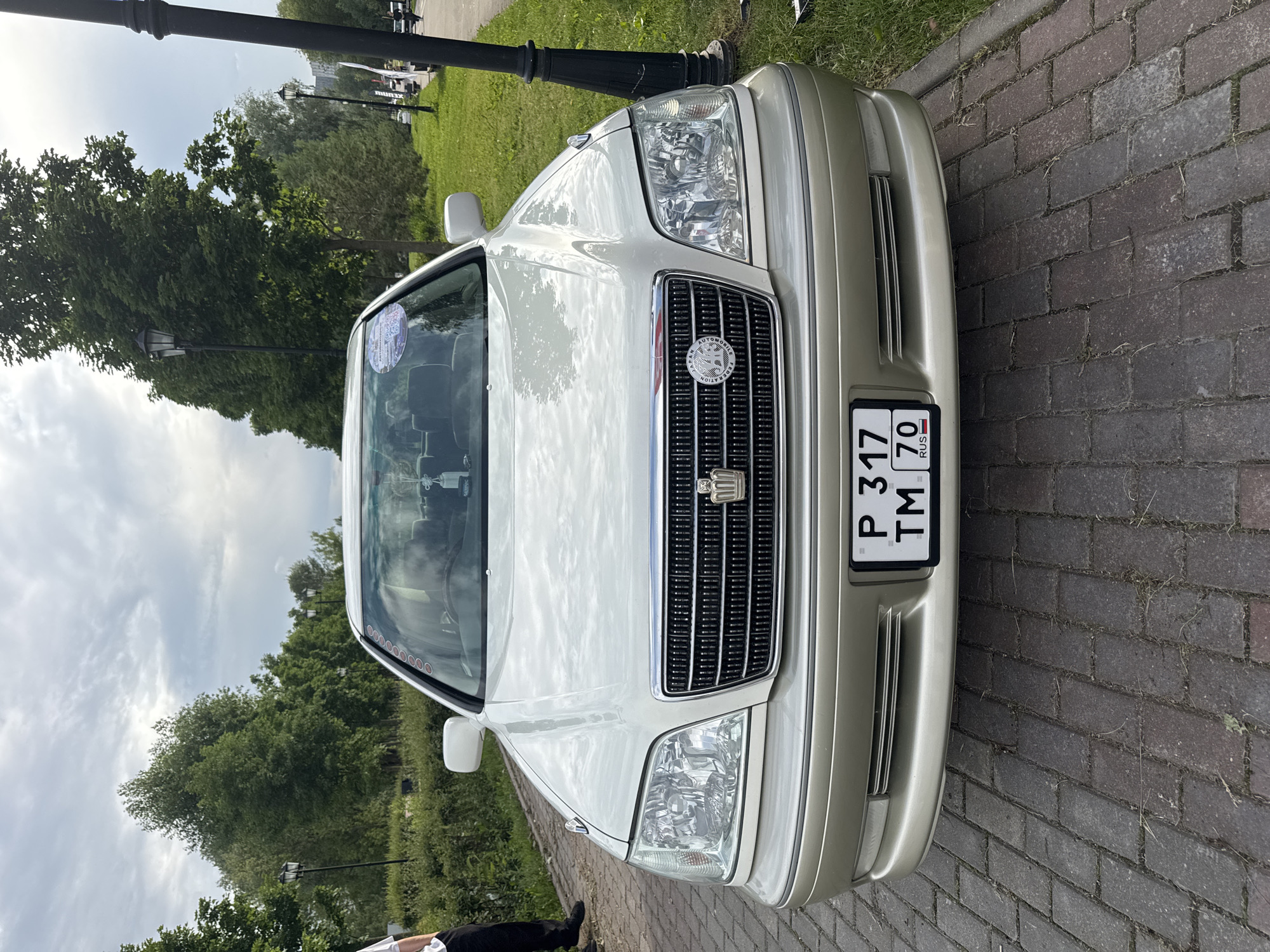 An eye-level, medium shot captures the front of a light-colored sedan parked on a brick pathway in a park-like setting. The car, identified by its license plate "P 317 TM 70," is the central focus. To the left of the car, the lower half of a person in black pants and a white shirt is visible, standing on the pathway. The background features lush green trees, some with streetlights, and a gray, cloudy sky. The environment suggests a city park in Moscow, Russia, likely on an overcast day.