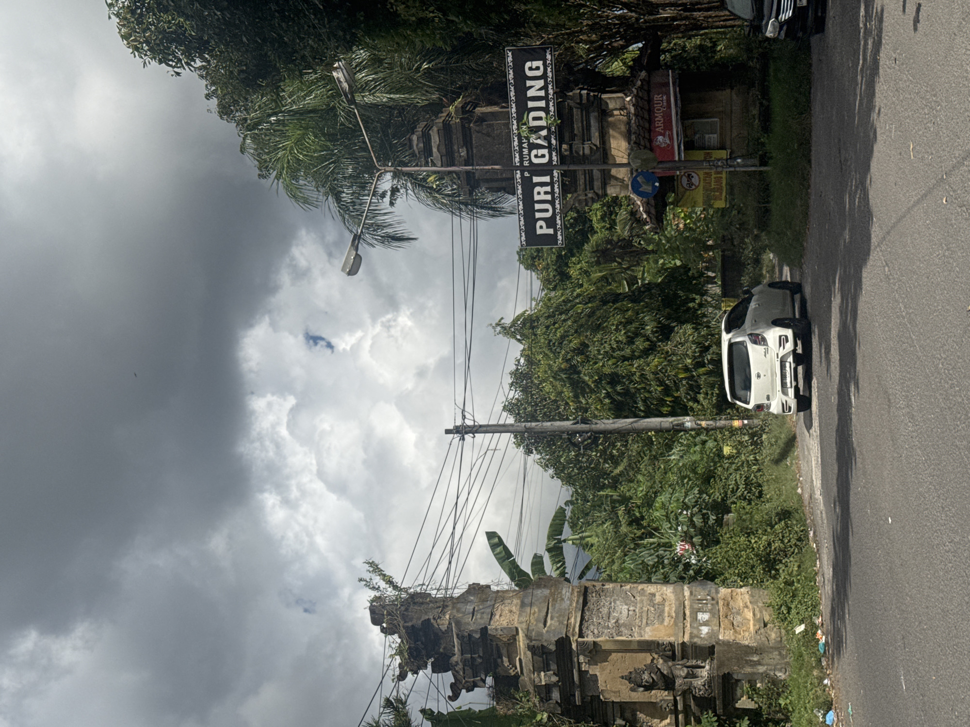 The image depicts a street scene in an unknown location, likely in a tropical or subtropical area, given the lush vegetation. A sign for "Puri Gading" is visible, indicating the name of a business or establishment in the area. The sky is cloudy and overcast, suggesting a recent or impending rain. A white car is parked on the side of the road. A stone structure, possibly an old gate or wall, is partially overgrown with vegetation. Power lines are visible overhead. The image appears to be taken from the side of the road, with the photographer facing toward the Puri Gading sign.