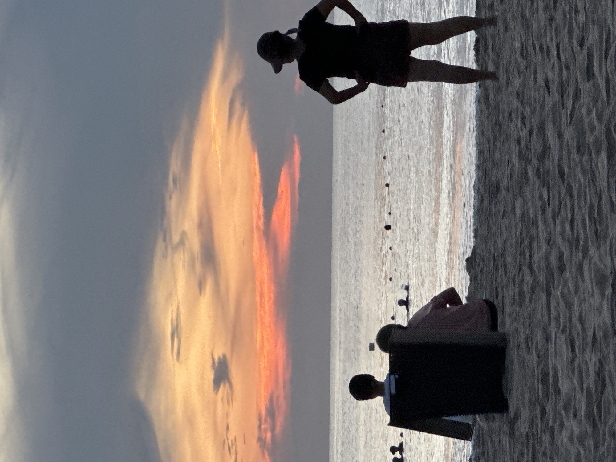 The image depicts a sunset scene at a beach, likely in Đà Nẵng, Vietnam, given the context. The sky is filled with vibrant orange and pink clouds, transitioning to darker grays at the top. The ocean is calm and reflects the colors of the sky.
In the foreground, on the sandy beach, are several individuals. To the left, two people are seated, one in a black lounge chair facing away from the camera and towards the ocean, and another person partially visible next to them, also seated. Further to the right, a person stands with their hands on their hips, wearing a dark t-shirt, shorts, and a hat, facing the ocean. In the middle ground, several other people are visible in the water, some appearing to be swimming or wading, marked by dark, rounded shapes. The beach itself is composed of sand with visible textures and ripples. The overall lighting suggests it is late afternoon or early evening.