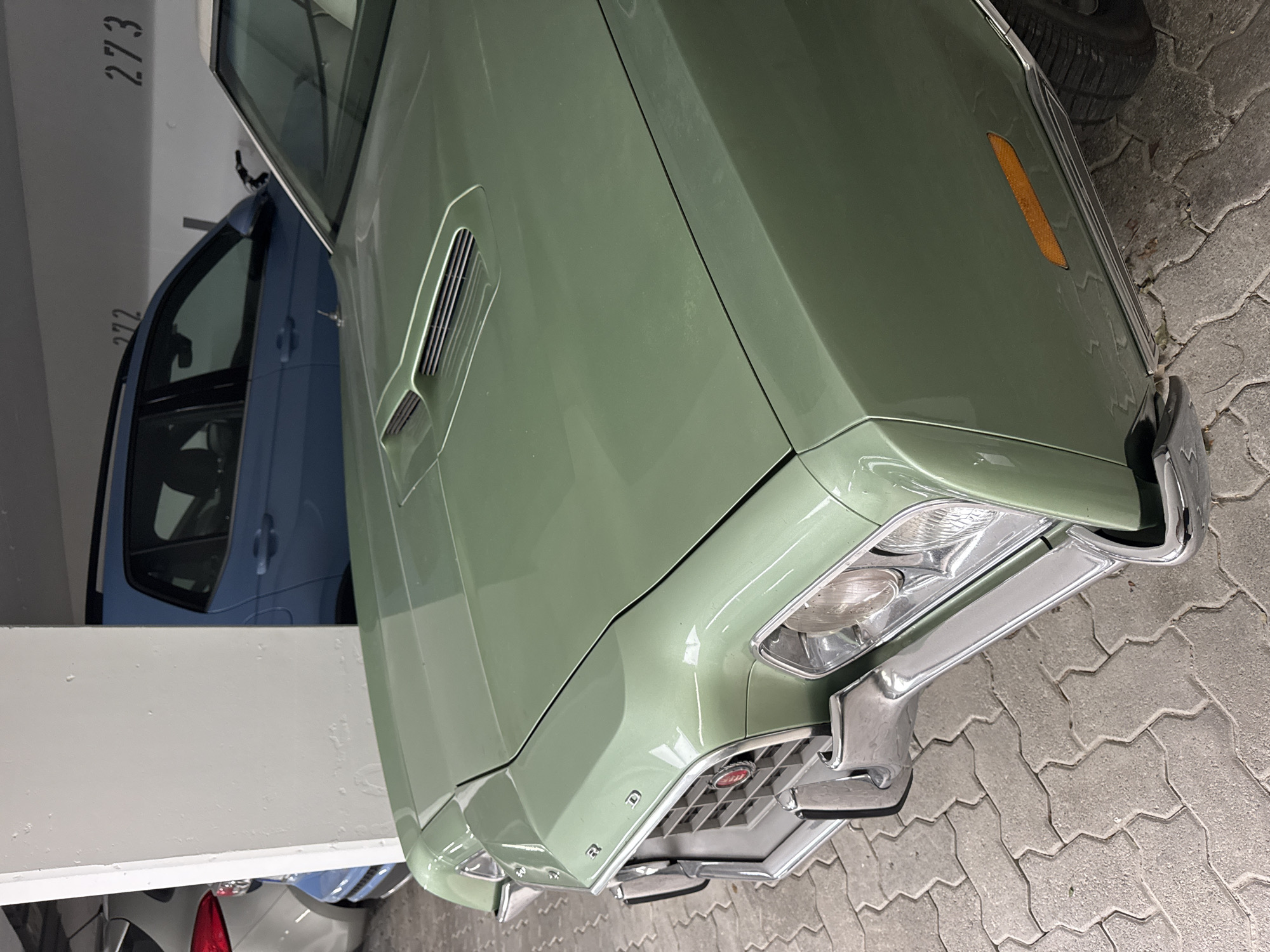 The image shows the front quarter of a light green 1970s Ford Gran Torino parked on interlocking pavers in an underground parking garage. The car has a prominent hood scoop and a chrome grille with a red circular emblem. Behind the Torino, a light blue SUV is parked, with the numbers "272" and "273" visible on the wall above it. To the left of the Torino, another vehicle, partially obscured, is parked. The environment is typical of an urban parking structure, with concrete walls and flooring. No people are visible in the image. The location is identified as Höhenkirchen-Siegertsbrunn, Germany.