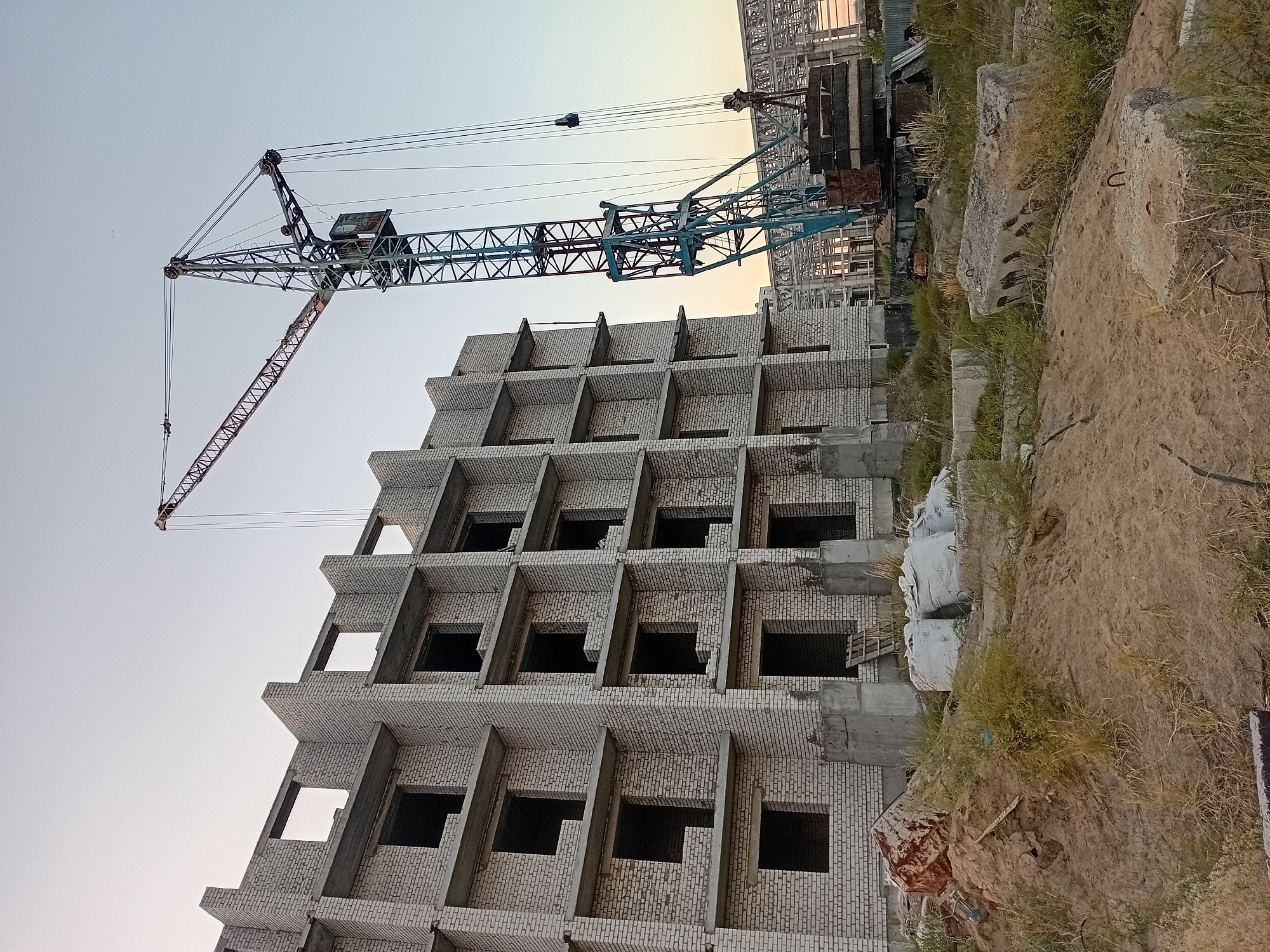 The image depicts an ongoing construction site in the city of Semey, Kazakhstan. A multi-story building, constructed with white bricks and concrete frames, is in the process of being built. Numerous window openings are visible on its facades, indicating unfinished interior spaces. Adjacent to the building stands a large construction crane, painted in a teal and red color scheme, with its boom extending over the structure. The surrounding area is a dirt and gravel lot, scattered with construction debris, including concrete blocks, sacks of material, and what appears to be a discarded metal object. Overgrown grass and weeds are present in patches throughout the site. In the background, other construction elements, such as metal frameworks, suggest further development. The sky is clear and pale, suggesting either early morning or late afternoon light. No people are visible in the frame.