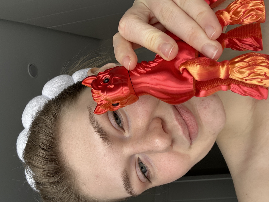 A close-up portrait in an indoor setting features a young adult female with fair skin and light brown hair, pulled back by a white, puffy headband. She exhibits a subtle smile towards the camera. Her right hand holds an articulated 3D-printed toy, resembling a horse or mythical creature, colored in a red-to-orange gradient. The toy is positioned near the right side of her face. The background is a uniformly blurred light grey wall, with a recessed ceiling light fixture visible in the upper portion. Visual information does not permit identification of a specific location within Kazan, Russia.