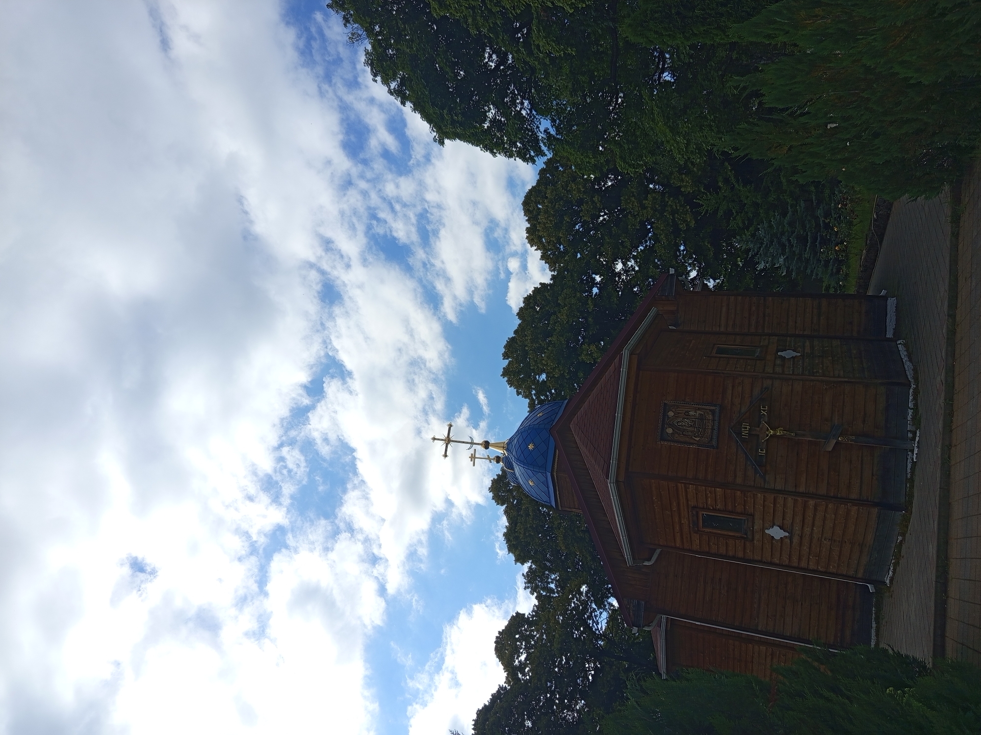 The image captures a wooden Orthodox church with a blue dome and a golden cross, set against a backdrop of lush green trees and a cloudy sky. The church, located in the city of Giaginskaya, Russia, has a rustic wooden facade with visible horizontal planks. A crucifixion scene is mounted on the exterior wall, featuring a golden crucifix with Jesus and inscriptions. The church has a red-tiled roof and several windows. The surrounding environment is characterized by dense foliage, including evergreen trees and bushes, and a paved walkway leading towards the church. The sky is a mix of blue and white clouds, suggesting a partly cloudy day. No people are visible in the image.