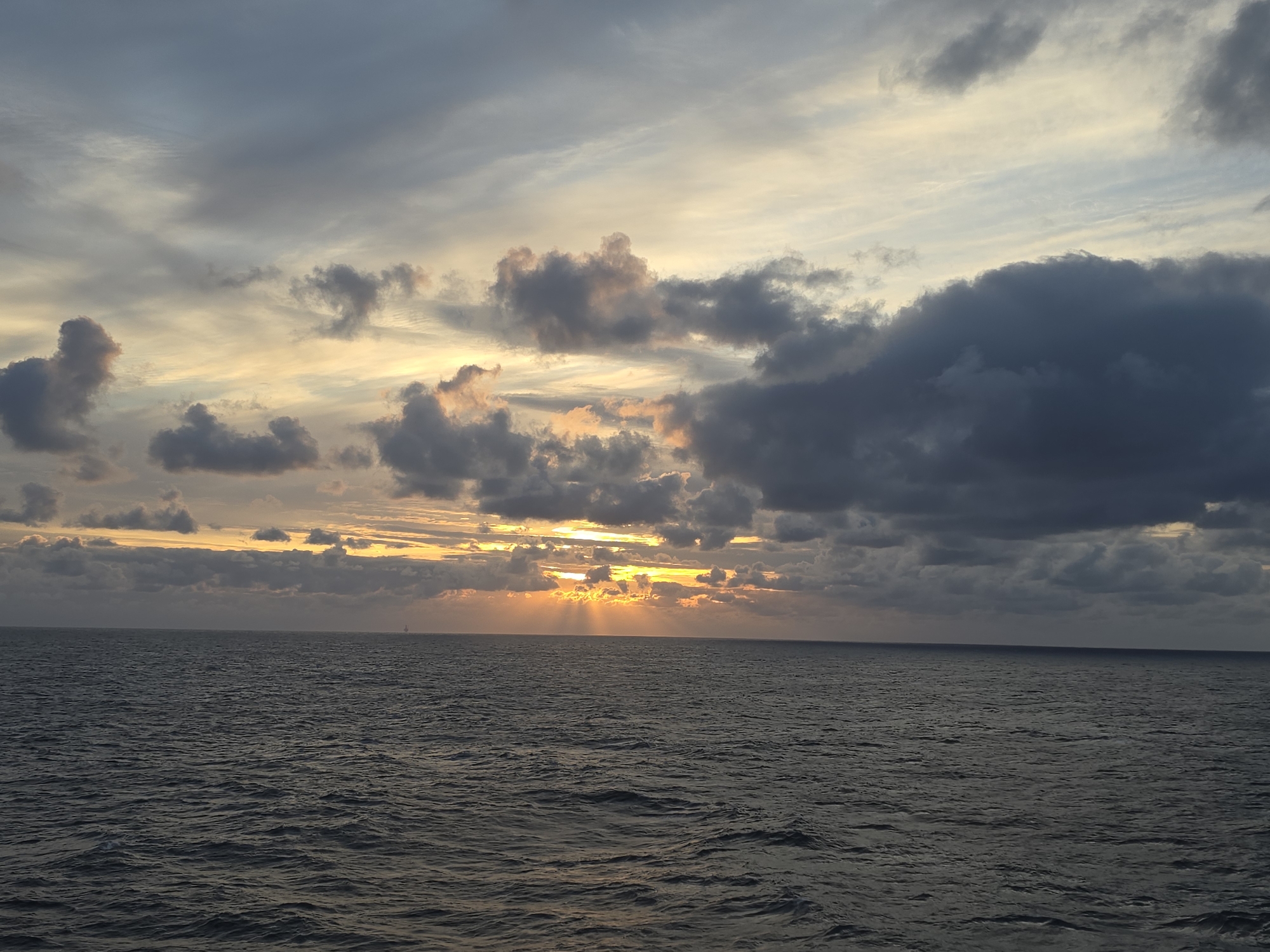 The image depicts a sea landscape at sunset. The sky is filled with a variety of clouds, ranging from light grey to dark grey, with patches of orange and yellow light peeking through. The sun's rays are visible as they break through the clouds and illuminate the water surface. The sea is dark and choppy, with small waves visible across the expanse. There is a single object on the horizon. The location appears to be a sea, city and date unknown.