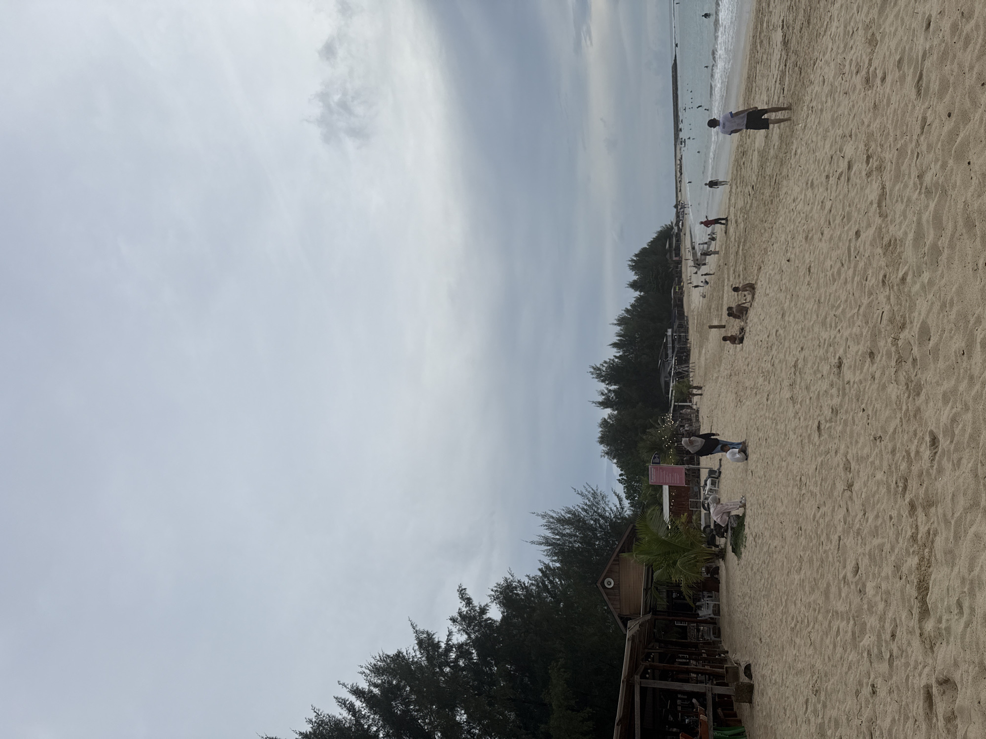 The image depicts a sandy beach with several people present. The sky is overcast with scattered clouds. On the left side of the image, there are some structures that appear to be wooden huts or stalls, with people visible inside some of them. Trees line the background behind these structures. Further down the beach, there are more people scattered along the sand and in the water. Some individuals are walking, while others are sitting or playing. The ocean is visible in the distance, with a breakwater or pier extending into the water on the far right. The sand is a light brown color, and there are footprints visible in some areas. The scene suggests a public beach with some amenities.