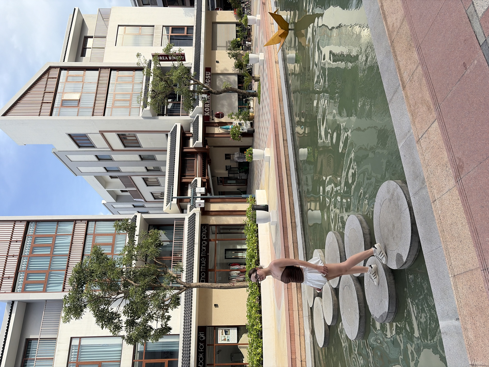 A young woman with dark hair, wearing a brown strapless top and a white skirt, stands on a large circular stepping stone in a shallow body of water. She is wearing glasses and white sneakers. The water reflects the surrounding buildings. In the background, there are modern, multi-story buildings with large windows and balconies, some adorned with wooden slats. Trees and potted plants are visible along the walkways and in front of the buildings. A yellow origami crane sculpture floats in the water to the right. Signs on the buildings indicate commercial establishments, with one reading "KOREA BINSU". The scene appears to be an outdoor public space within a planned urban development in Nghĩa Trụ Commune, Vietnam, possibly a commercial or residential complex. The lighting suggests it is daytime, with clear skies and some shadows.