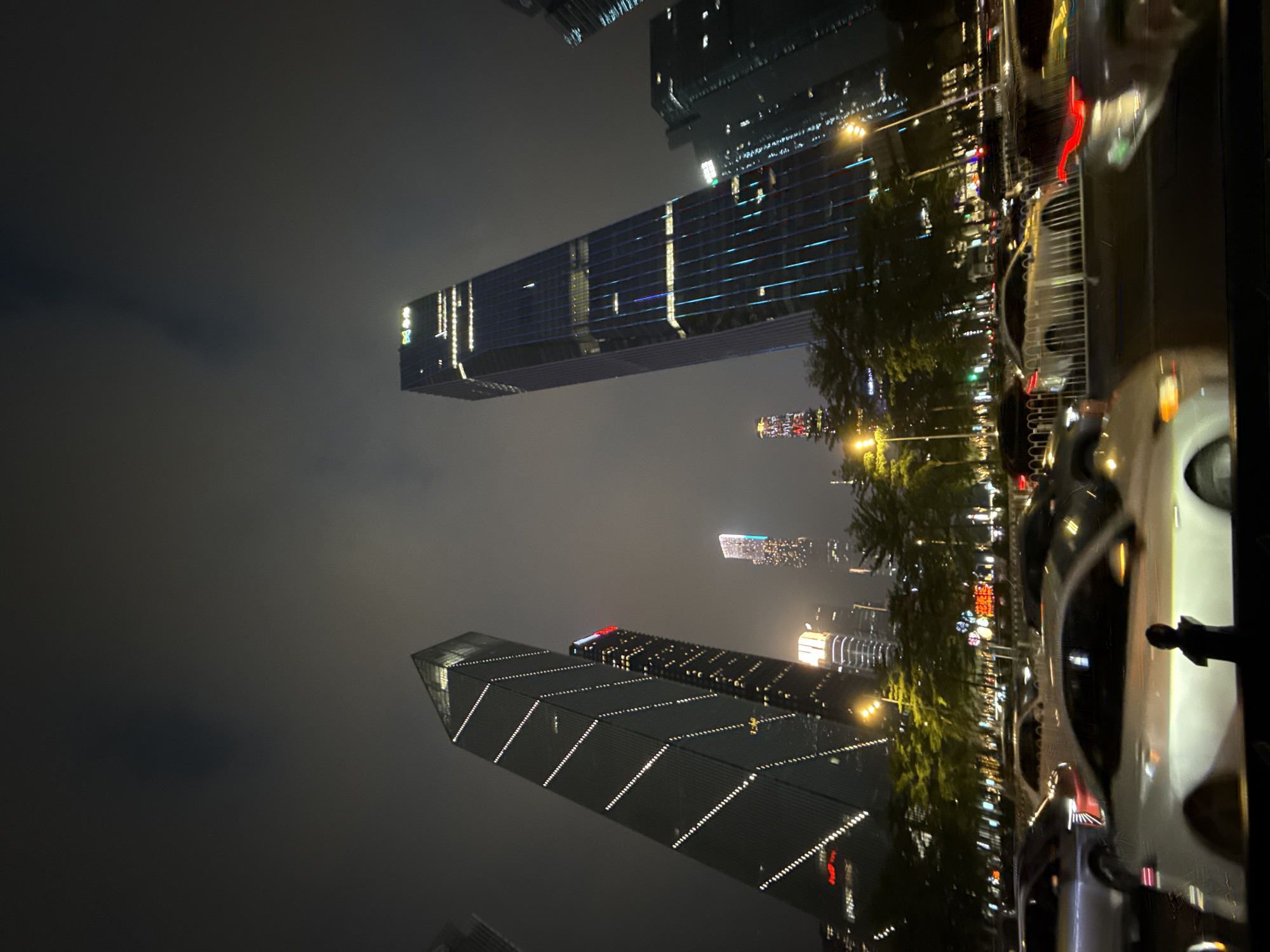 The image shows a nighttime street scene in Tianhe District, China. The view is angled upwards towards modern skyscrapers illuminated with lights. On the right side of the frame, there are parked cars lining the street. Trees are visible in the middle ground along the street. The sky is dark and cloudy.