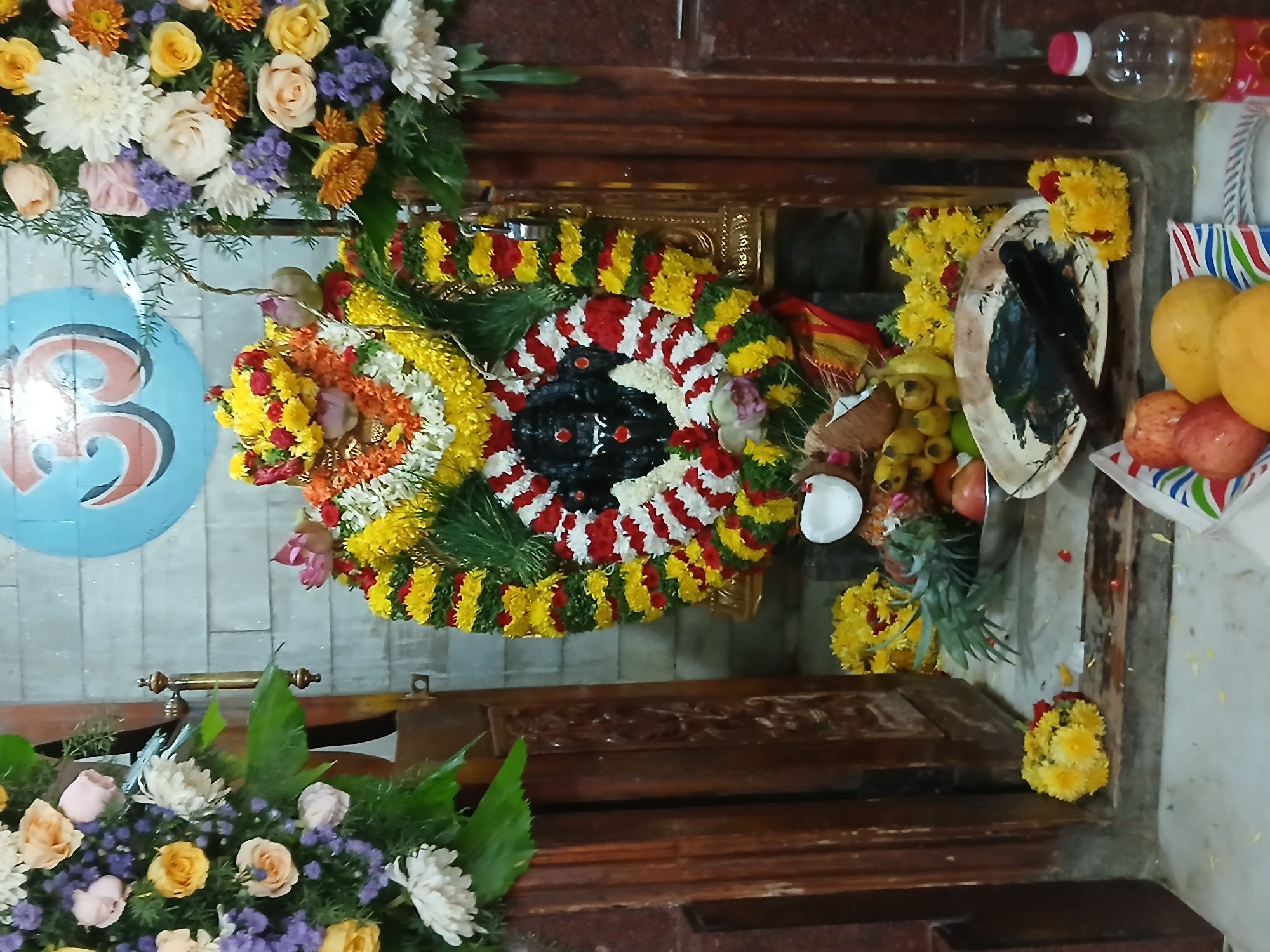 The image shows a Hindu shrine, likely within a temple or private home. The central focus is a deity figure adorned with garlands of yellow, red, and white flowers. Various offerings, including fruit (mangoes, pomegranates) and coconuts, are arranged around the deity. A decorative plate with what appears to be a painted or carved image stands next to the offerings.
The shrine is housed within a wooden structure with ornate detailing. Flowers are prominent around the scene. A blue roundel with red numbers adorns the wall. A plastic bottle sits above the shrine.
Without further evidence, it is impossible to verify the location as Bengaluru, India.