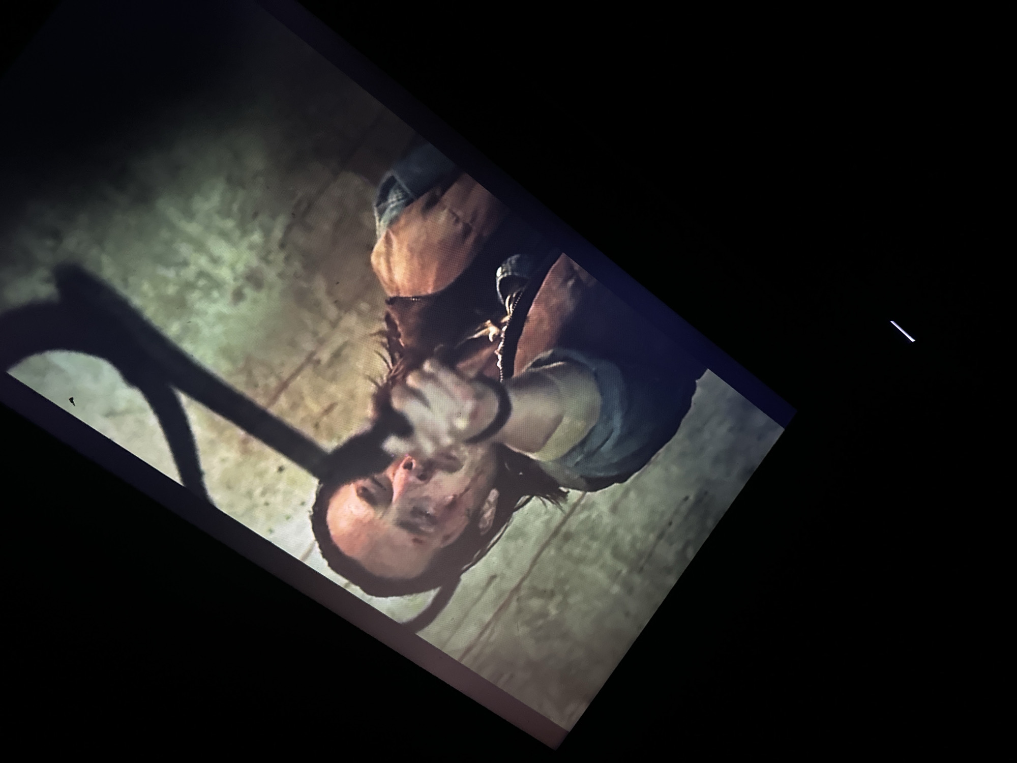 This dimly lit, rotated image captures an intense, action-oriented scene, likely a screengrab from a film or television show. The central subject is a male individual, appearing to be in his 30s or 40s, with dark, shoulder-length hair and a beard or stubble. His face is visibly dirty and marked with what appear to be cuts or blood, suggesting he has been in a struggle. His expression is strained, conveying a mix of pain, fear, or fierce determination, as he looks directly or nearly directly towards the camera.
He is dressed in a dark blue or denim-colored long-sleeved shirt underneath a reddish-brown vest or jacket. A necklace or pendant is visible around his neck. His right arm is extended forward, pointing an indistinct dark object, possibly a weapon like a knife or the barrel of a gun, directly at the viewer's perspective. The angle of the shot suggests the camera is looking up at him, enhancing the confrontational and immersive feel of the moment.
The background is dark and indistinct, featuring a textured surface that resembles a dirty, rough floor or wall, possibly made of stone or concrete, in muted green-grey tones. To the left, a dark, curved object, possibly part of a railing, pipe, or structural element, casts a prominent shadow. The overall lighting is low and artificial, contributing to a gritty and suspenseful atmosphere. No text is visible in the image.
The scene depicts a moment of high tension or conflict, with the man seemingly in mid-action, possibly defending himself, attacking, or caught in a perilous situation. The image itself provides no visual cues to connect it with the specified location of Saint-Leu-d'Esserent, France.