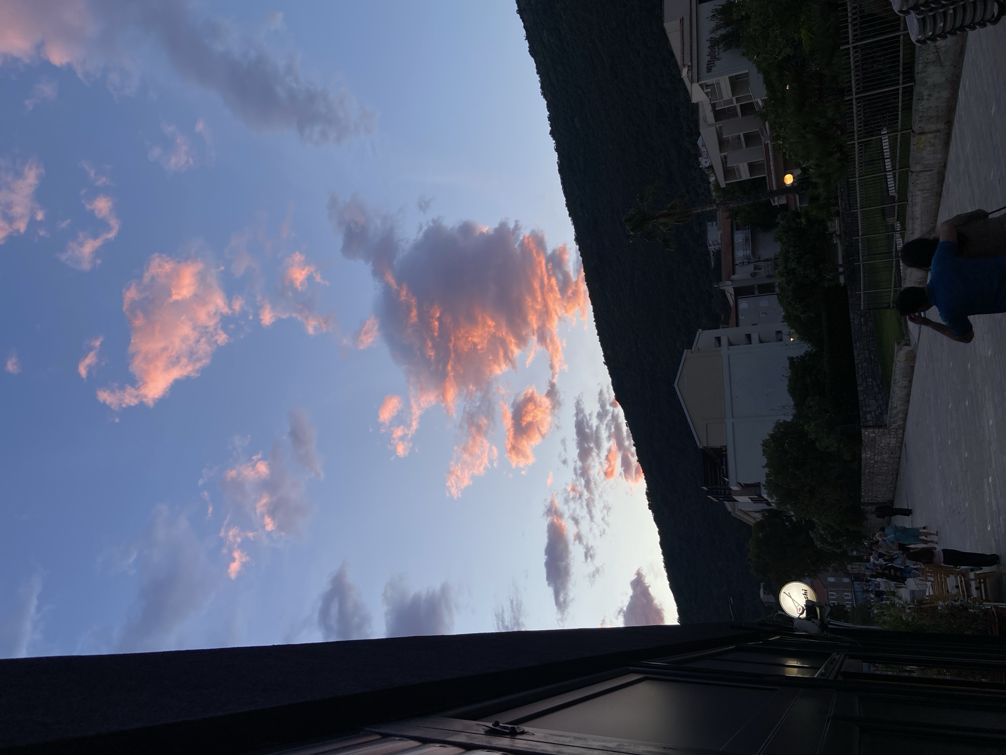 The image shows a view in Petrovac centar, Montenegro, taken from a partially visible rooftop or balcony. A mountain occupies the central right of the frame. To the right of the mountain are buildings with trees and a street with people. Above the mountain is a vibrant, pink and blue sunset with a few clouds. The people appear to be socializing and walking in the street. A restaurant sign is visible.