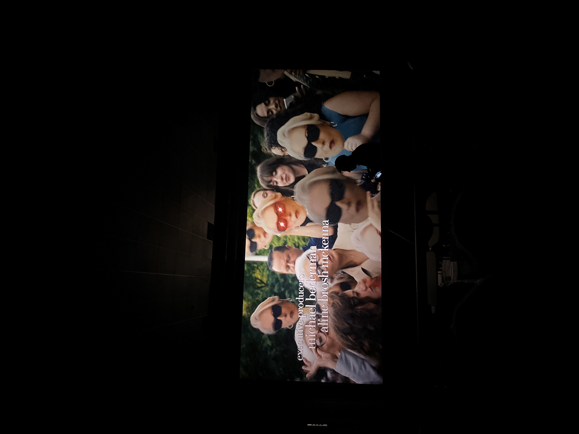 The image is a photograph taken of a movie screen in a dark cinema. The screen displays the end credits of a film, with the names "executive producers michael bederman aline brosh mckenna" superimposed over a crowd of people wearing sunglasses. The people are dressed in various styles and are looking at the camera. The cinema setting is suggested by the darkness surrounding the screen and the presence of what appears to be a chair back in the foreground. There is no evidence to suggest the location is Puebla, Mexico.
