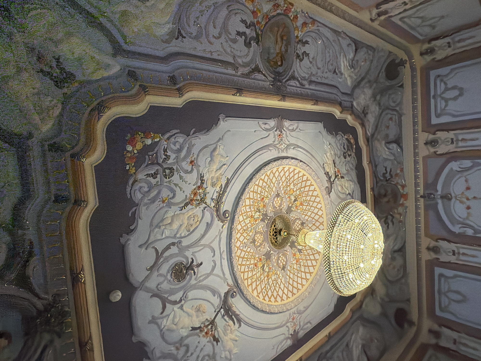 The image shows an ornate ceiling with a chandelier in the interior of a building. The ceiling is decorated with elaborate plasterwork and painted murals, featuring cherubic figures, floral motifs, and what appears to be a pastoral scene with trees and foliage. A large, crystal chandelier hangs from the center of a decorative medallion on the ceiling. The building's interior walls are also visible in the lower part of the image, adorned with decorative molding and painted panels. The style suggests a grand and historic building. The location is identified as the Nizhny Novgorod State Academic Drama Theatre named after Maxim Gorky, in Nizhny Novgorod, Russia, specifically the ceiling of the auditorium. There are no people visible in the image, and the focus is on the architecture and decoration of the space.