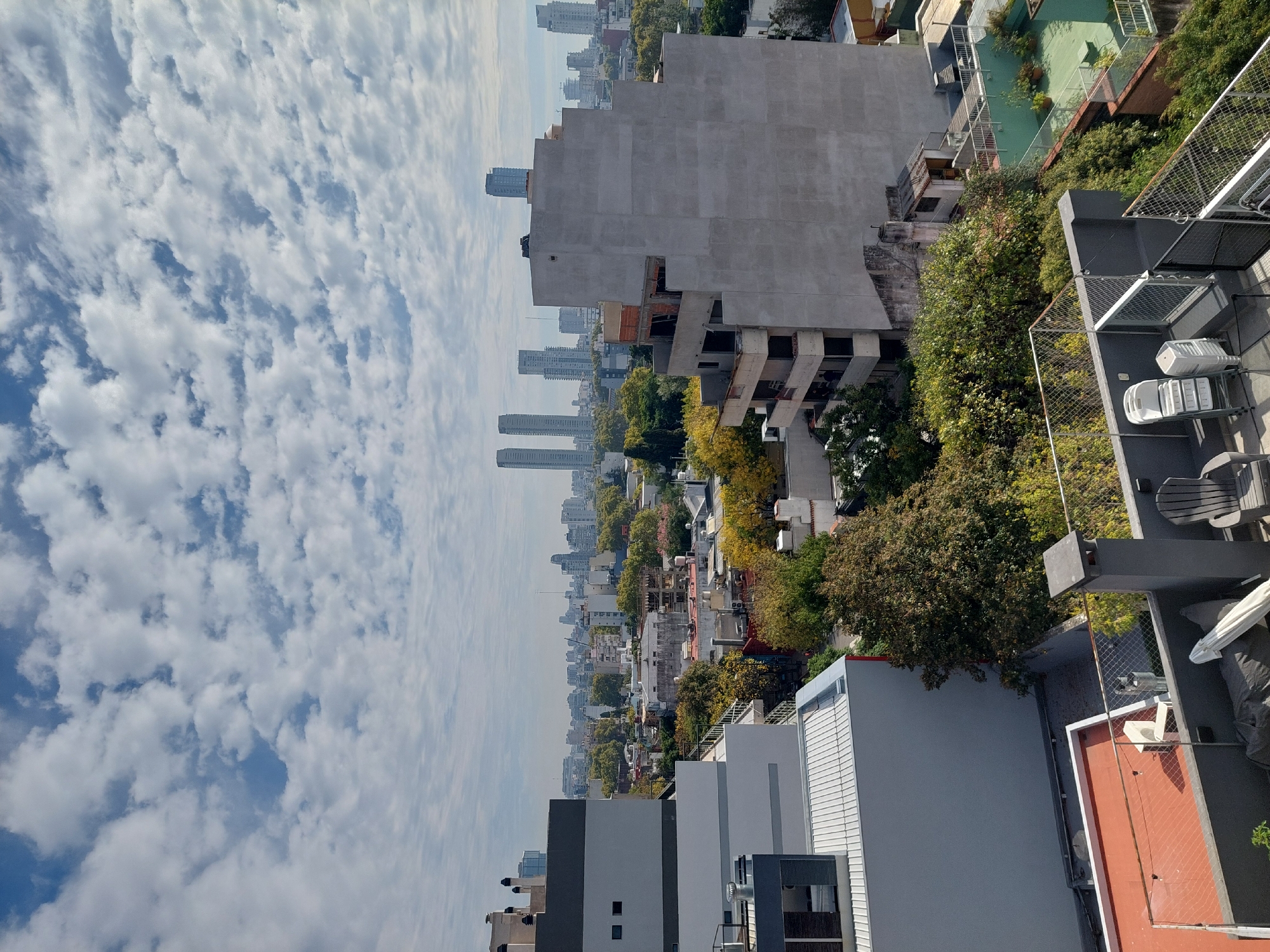 The image shows a cityscape in Buenos Aires, Argentina, likely taken from a high vantage point. In the foreground, there are rooftops of buildings, including some with terraces. One terrace has what appears to be patio furniture. A large unfinished concrete building stands prominently in the middle ground. In the distance, a row of high-rise buildings and skyscrapers lines the horizon. The sky is filled with numerous puffy white clouds. The scene includes residential and commercial buildings, trees, and the sky. No people are visible in the image.