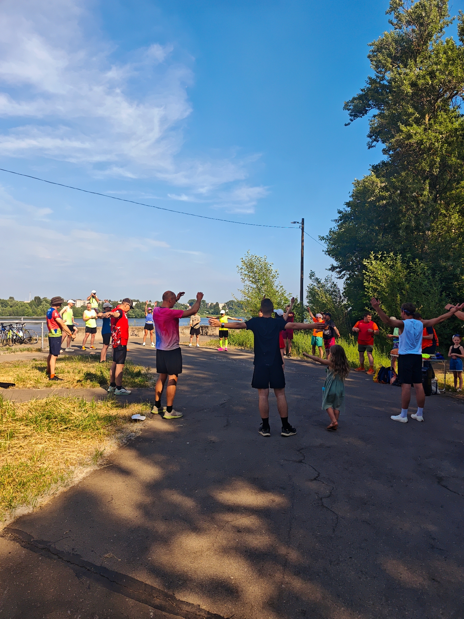 The image depicts an outdoor group exercise class taking place on an asphalt road next to a grassy area. A body of water, likely the Volga River, is visible in the background, with trees and buildings lining the opposite bank, suggesting a riverside urban setting, consistent with the city of Kimry, Russia. The sky is blue with scattered white clouds.
A diverse group of approximately 15 individuals, including adults and one young child, are engaged in stretching or warm-up exercises. Most are facing away from the camera, with their arms extended. They are dressed in athletic wear, with bright colors being common. Notable individuals include a person in a pink and white tie-dye shirt in the foreground, and a person in a light blue tank top to their right. A young girl in a green dress is also participating, positioned slightly ahead of the main group.
To the left of the scene, several bicycles are parked near the grassy verge. The environment appears to be a park or recreational area. Shadows from trees are cast across the asphalt, indicating a sunny day.