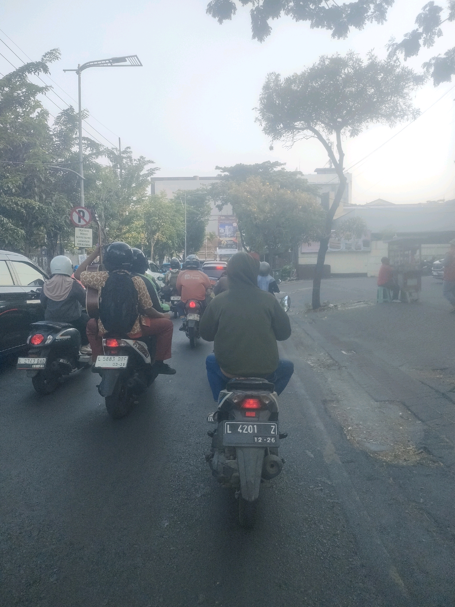 The image depicts a traffic jam on a street in Surabaya, Indonesia, characterized by heavy fog and low visibility. Several motorcycles and their riders are visible, navigating through the congested road. One person is riding a motorcycle while carrying a guitar. The environment suggests an urban setting with streetlights, trees, and buildings lining the road. The time of day appears to be morning or evening, given the lighting conditions and the fog. The presence of license plates indicates that the vehicles are registered and operating within Indonesia.