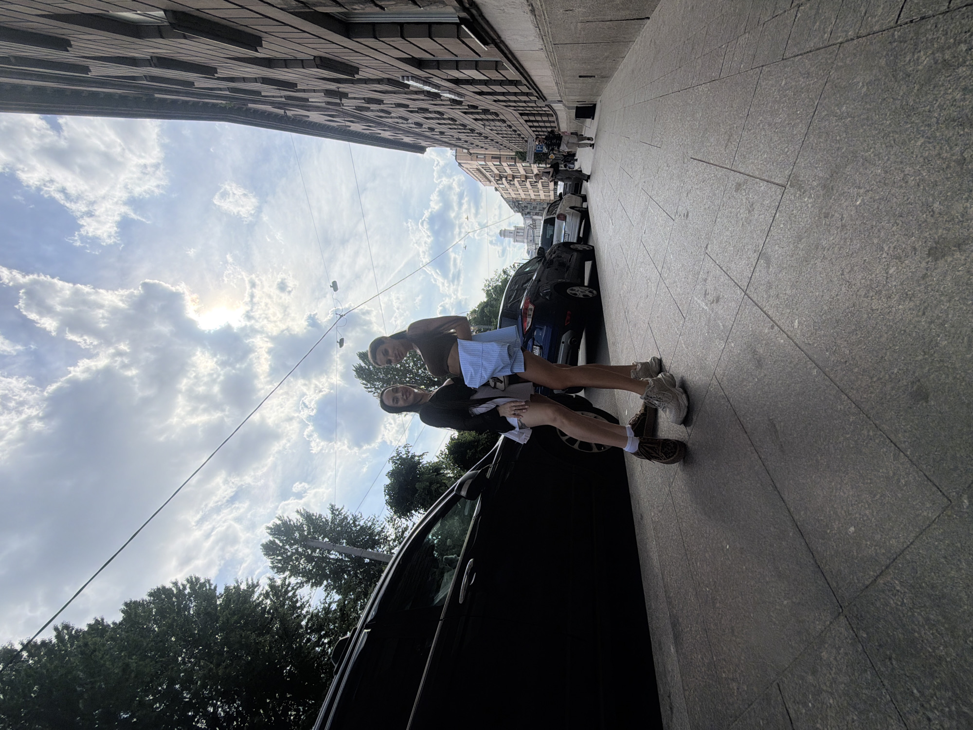This image depicts a sunlit urban street in Saint Petersburg, Russia, on a partly cloudy day. In the foreground, two young women are posing on a grey tiled sidewalk. The woman on the left, with long dark hair, wears a black blazer over a white garment, a short light-colored skirt, white socks, and brown lace-up shoes; she smiles towards the camera. The woman on the right, also with long dark hair pulled back, wears a dark brown crop top and light blue striped shorts, with light-colored chunky sneakers; she smiles with her left hand resting on her stomach.
A black SUV is parked parallel to the sidewalk immediately behind the women. Further down the street, additional parked vehicles include a blue SUV, a silver sedan, and a white van. Several pedestrians are visible in the mid-ground, including a group of three (two women and one man) on the sidewalk. Overhead utility wires are strung across the street.
The right side of the street is lined with a multi-story building featuring a dark, stone-paneled facade with numerous rectangular windows. Trees are visible along the left side of the street. In the far distance, the distinctive golden dome and colonnade of Saint Isaac's Cathedral are clearly discernible against a sky with scattered white clouds and visible sunlight.
The presence of Saint Isaac's Cathedral in the background confirms the location as the historic center of Saint Petersburg, Russia, likely a street leading towards the cathedral, such as Bolshaya Morskaya Ulitsa.