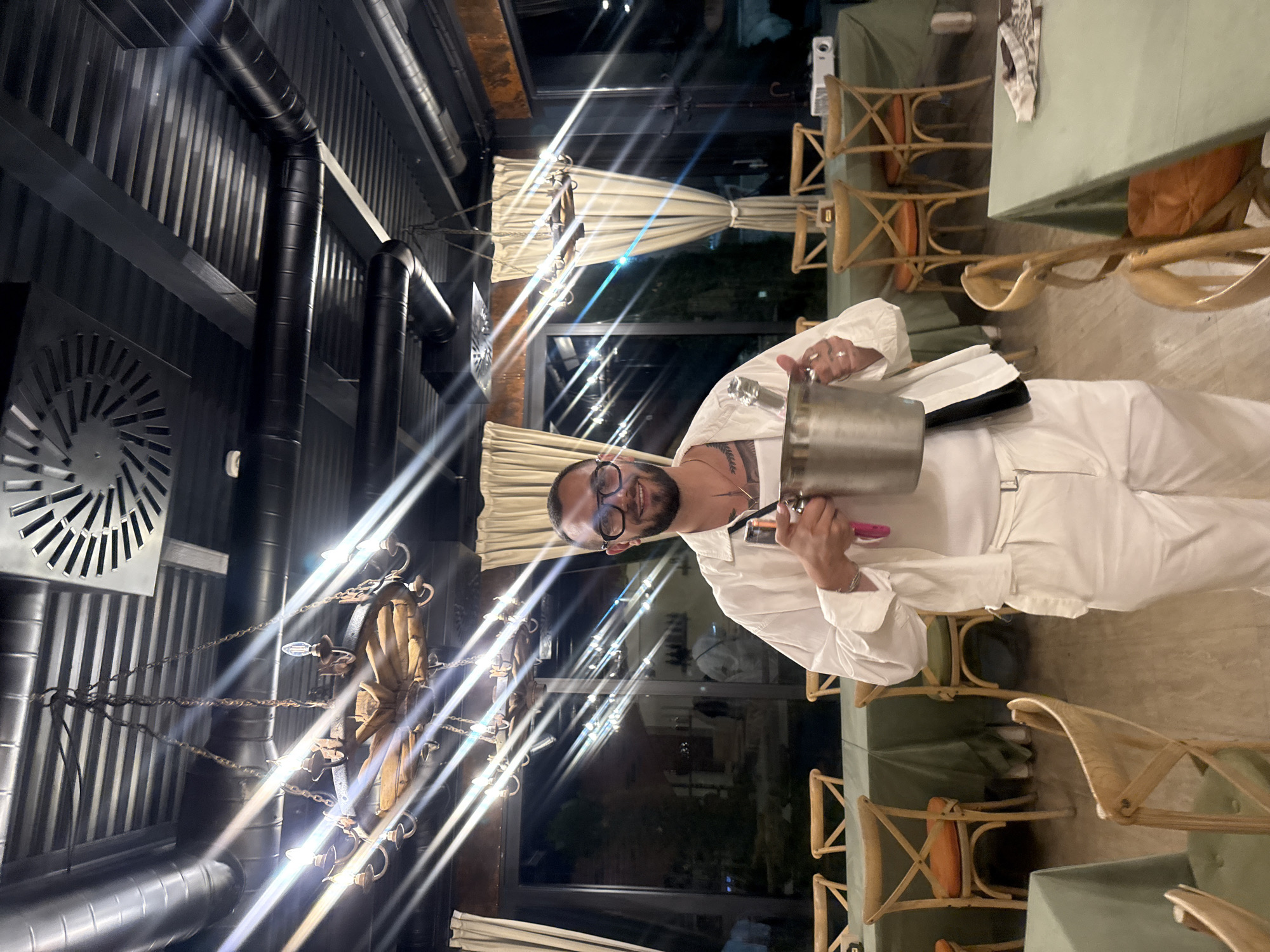 The image depicts a man standing in a restaurant dining area, possibly located in Sasunik, Armenia. He is wearing an all-white outfit, including a collared shirt and pants, and large-framed glasses. He is holding a silver bucket containing a bottle, likely for chilling drinks. The restaurant features numerous wooden chairs with orange or green cushions around tables covered in olive-green tablecloths. The ceiling is industrial, with exposed ducts and a large chandelier. Large windows in the background offer a view of an outdoor area with trees and lights, suggesting it is evening. The man is smiling and looking directly at the camera, holding the bucket with his right hand and what appears to be a bottle opener or similar tool in his left. The overall impression is one of a well-dressed individual presenting a beverage service in a modern, dimly lit restaurant setting.