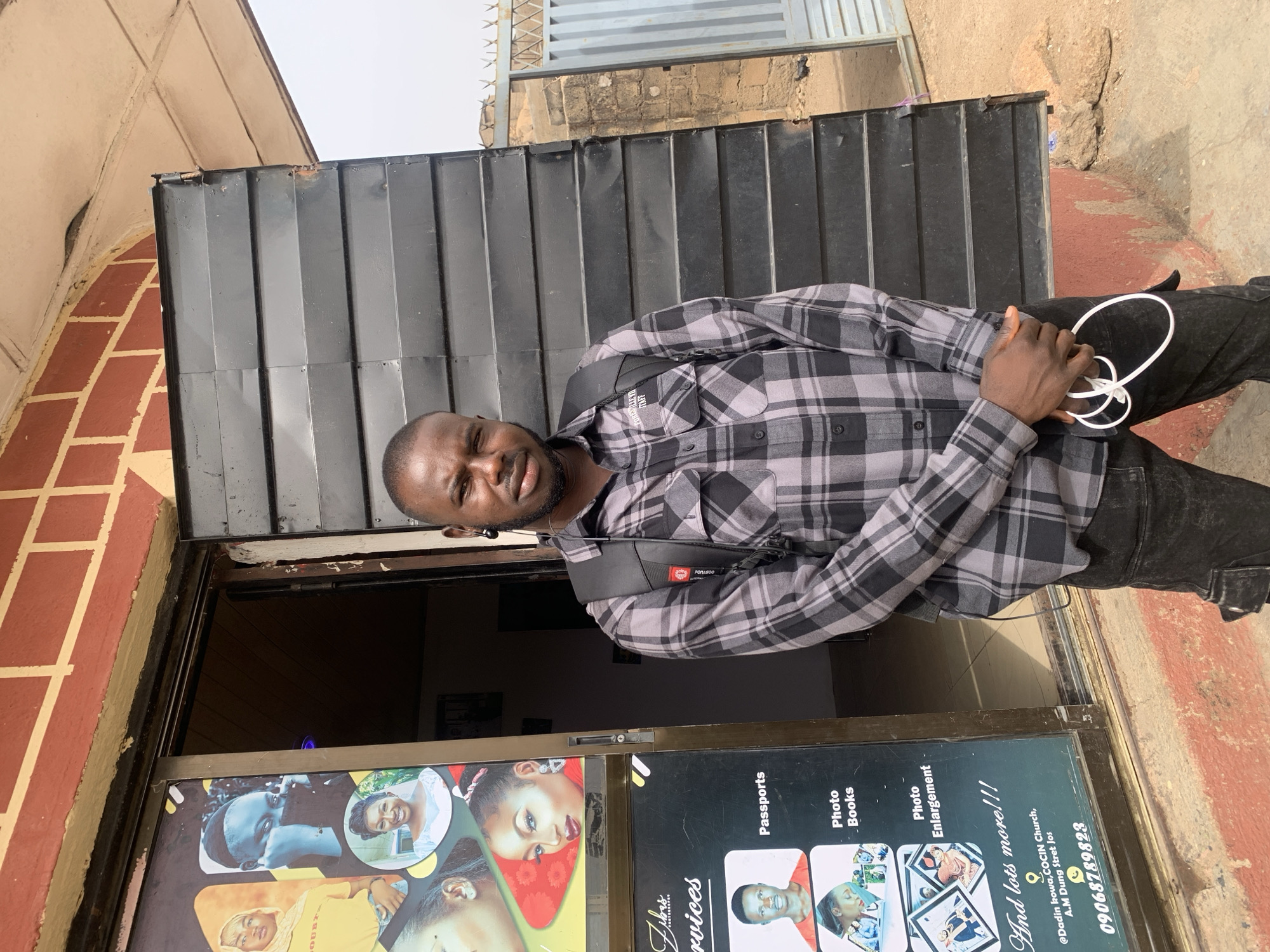 A man stands in a doorway, looking directly at the camera, in an outdoor setting during daytime in Jos, Nigeria. He is positioned slightly off-center to the right of the frame, wearing a grey and black plaid long-sleeved shirt, dark trousers, and a black shoulder bag. His left hand holds a white coiled cable, possibly a charging cable or headphones. He has short dark hair and a beard or stubble, with a neutral to slightly pleasant expression.
Behind the man is a black, vertically corrugated metal shutter or door. To his left, within the doorway, is a shop window displaying various photographic examples and services offered. The visible text on the window advertises "Passports," "Photo Books," "Photo Enlargement," and "And lots more!!!" It also identifies the business as "Zibas Services" (partially visible) and provides a location cue: "@Dadin kowa, COCIN Church, A.M Dung Stret Jos," along with a phone number: "09068789823." To the man's right, a reddish-brown brick wall is visible, suggesting the exterior of a building. The lighting is bright and even, indicating clear weather.