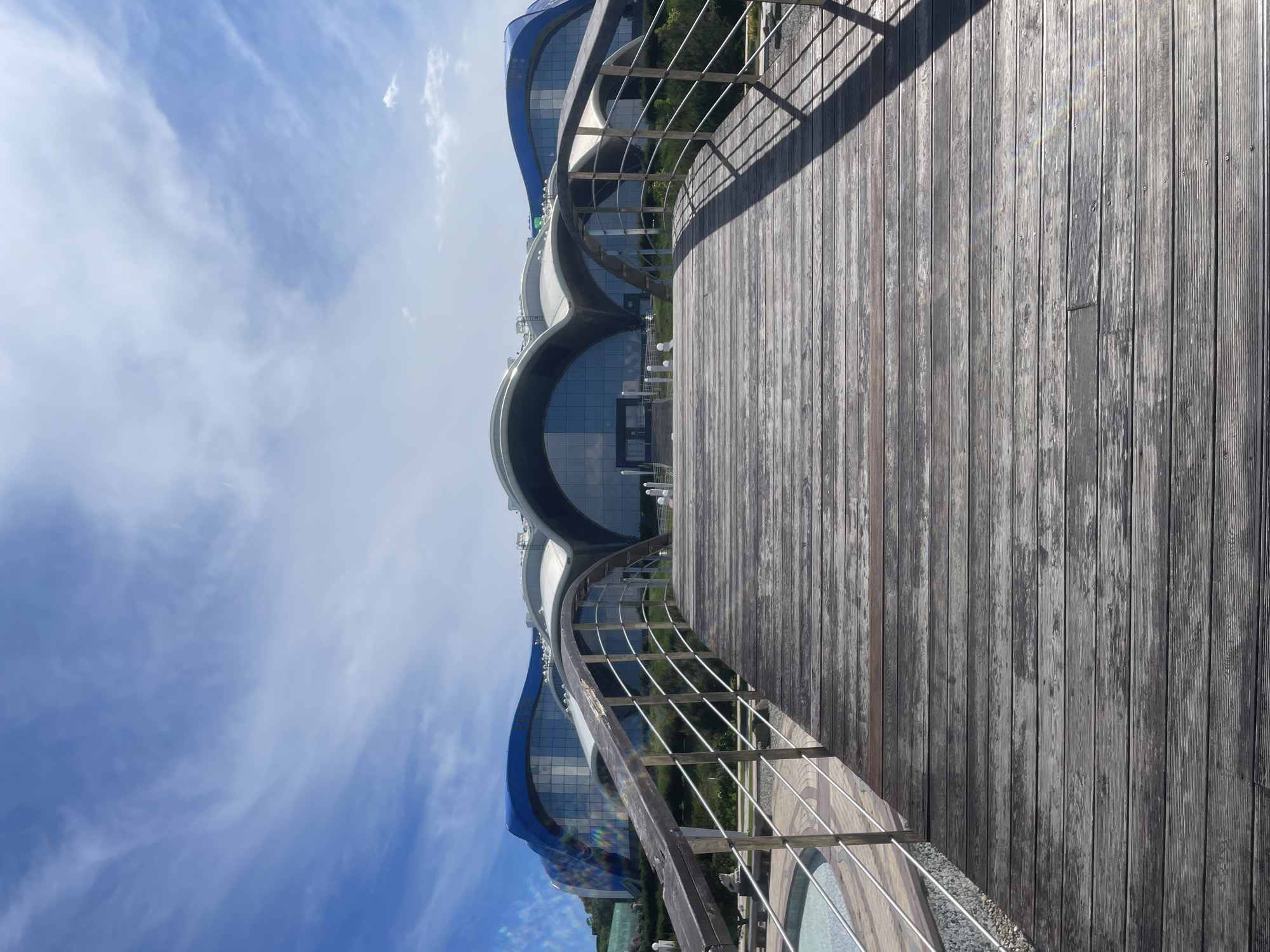 The image shows a wide wooden bridge with metal railings leading towards a modern, curved, multi-story building with a blue and silver facade. The building has large glass windows and appears to be an entertainment or educational facility. The sky is blue with scattered white clouds. The foreground of the image is dominated by the weathered wooden planks of the bridge. To the left of the bridge, a circular pool is partially visible. The scene is an outdoor, daytime view. The location is the Far Eastern Federal University campus on Russky Island, Vladivostok, Russia, likely depicting the Primorsky Aquarium.