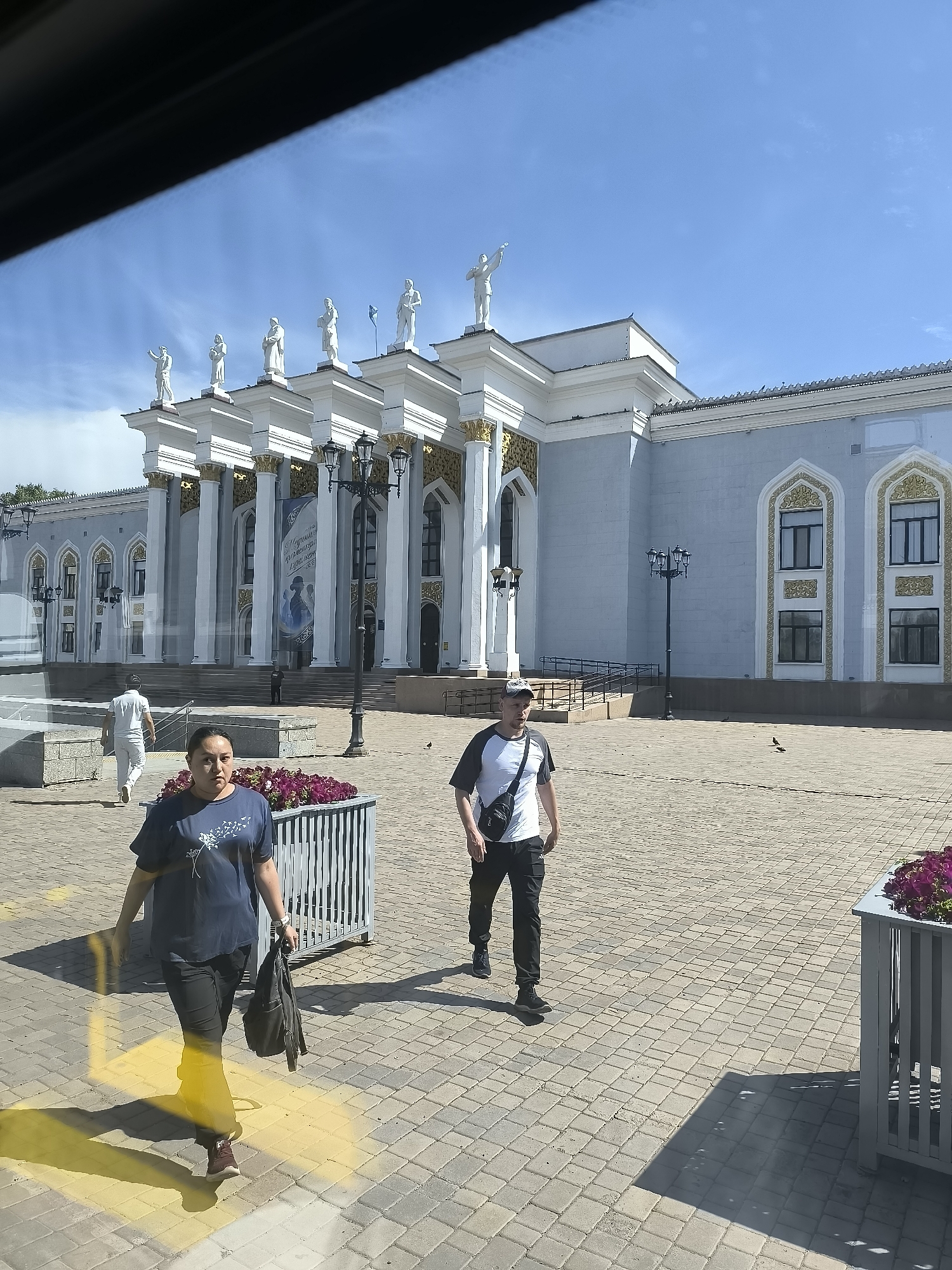 In Qarağandı, Kazakhstan, the city administration building is prominently featured, distinguished by its light blue facade, numerous white columns topped with golden embellishments, and a row of white statues on its roofline. The ground in front of the building is paved with light-colored square stones. Two individuals are walking in the foreground. A woman in a dark blue t-shirt with a dandelion print and dark pants walks towards the viewer, carrying a dark bag in her left hand. To her right and slightly behind her, a man in a black and white t-shirt, black pants, and a cap walks away from the viewer, with a black bag slung across his chest. In the middle ground, a person in a white t-shirt and white pants walks away from the viewer, towards the building. Planters filled with purple flowers are visible in the foreground and to the right of the image. Lampposts are also present. The sky is clear and blue, indicating a sunny day.