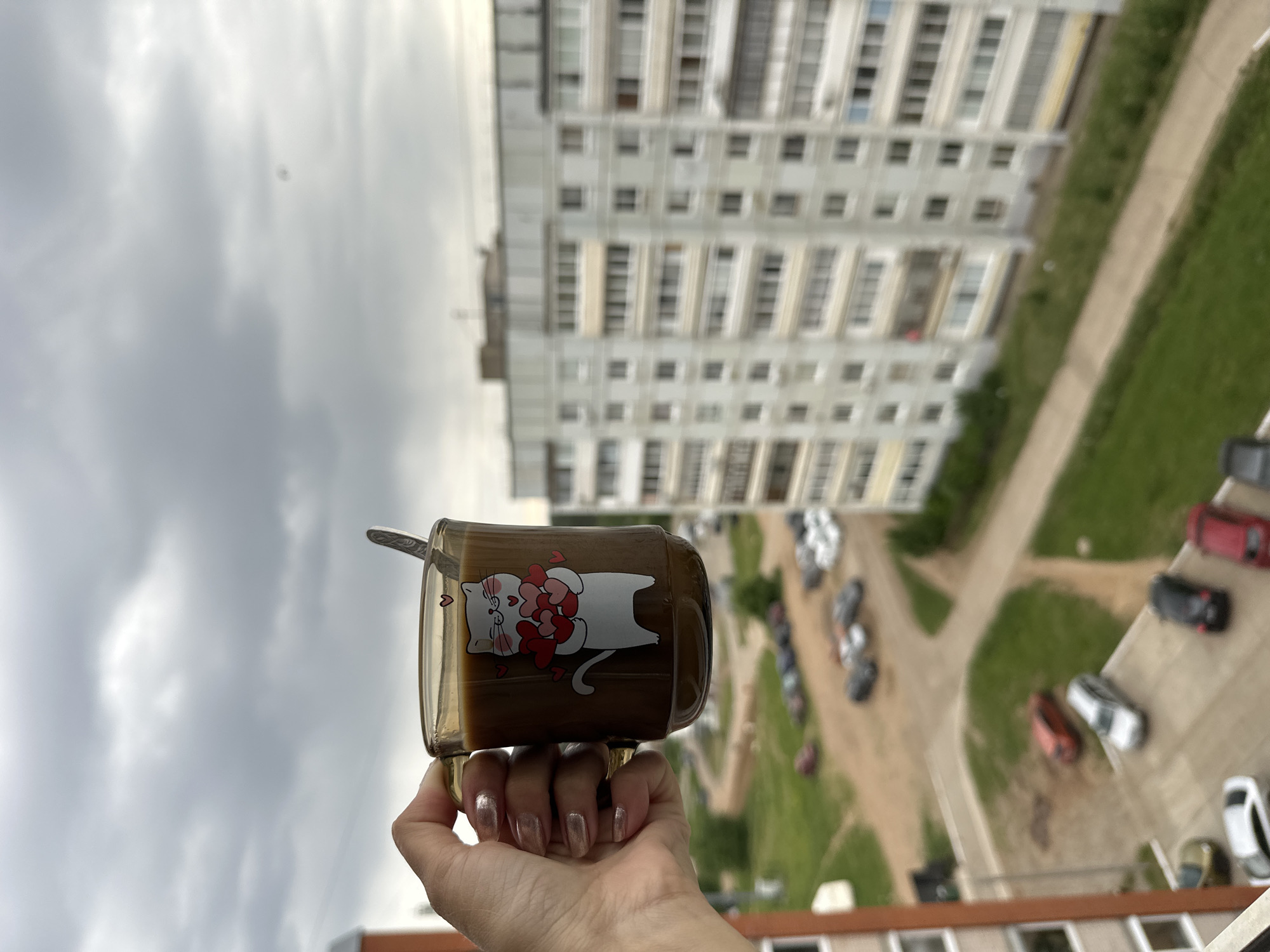 A close-up, eye-level shot captures a hand holding a glass mug against the backdrop of a residential area in Chaikovsky, Russia. The image is taken from a high vantage point, possibly a balcony or window, looking down and across.
In the foreground, a person's hand, adorned with metallic rose-gold nail polish, firmly grasps a clear glass mug. The mug contains a dark, creamy beverage, likely coffee or a similar drink, and a silver spoon rests inside. A whimsical cartoon cat design is printed on the mug, depicting a white cat with several red hearts floating above its head and paws.
The midground reveals a residential scene with a parking area and green spaces. Numerous cars are parked, some on paved lots and others on unpaved ground or grass. Dirt or gravel paths weave through grassy sections.
Dominating the background is a large, multi-story apartment building, light in color with many windows. Its architecture suggests a typical residential block. The sky above is overcast with visible clouds, indicating a cloudy daytime. No specific text is visible apart from the cartoon design on the mug. The overall atmosphere is mundane, capturing a quiet moment of enjoying a drink with a view of a city neighborhood.