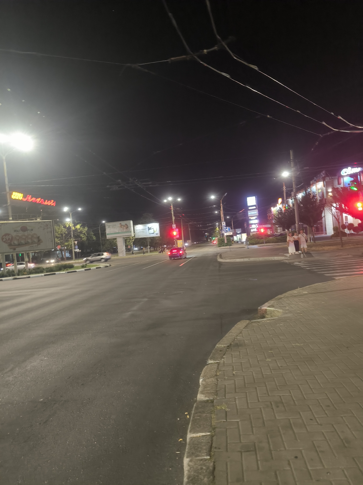 This is a night-time, ground-level, wide-angle photograph of a city intersection in Mykolaiv, Ukraine. The scene is illuminated by streetlights and the lights from businesses and vehicles.
In the foreground, a paved sidewalk with a herringbone pattern is visible on the right side of the image. The sidewalk is bordered by a curb and a street.
The street itself is a multi-lane road with a visible lane marking and some scattered debris. Several cars are present on the road, including a dark-colored sedan on the right side of the frame with its taillights illuminated and a light-colored sedan in the distance in the center of the frame.
Across the street, on the left side of the image, there are several large billboards. One billboard has the number "299" prominently displayed along with images of what appear to be fruits or vegetables. Another billboard has text in what appears to be Cyrillic script.
Further down the street, there are buildings with illuminated signs. One sign on the right side of the image appears to be for a business called "Cineplex" or a similar establishment, with neon lights. There are also traffic lights visible, with a red light illuminated at the intersection on the right.
On the crosswalk in the middle of the intersection, three individuals are standing, two women and one man, all dressed in light-colored clothing. They appear to be waiting to cross the street.
Overhead, power lines crisscross the dark night sky. The overall impression is of a typical urban intersection at night, with ongoing traffic and pedestrian activity.