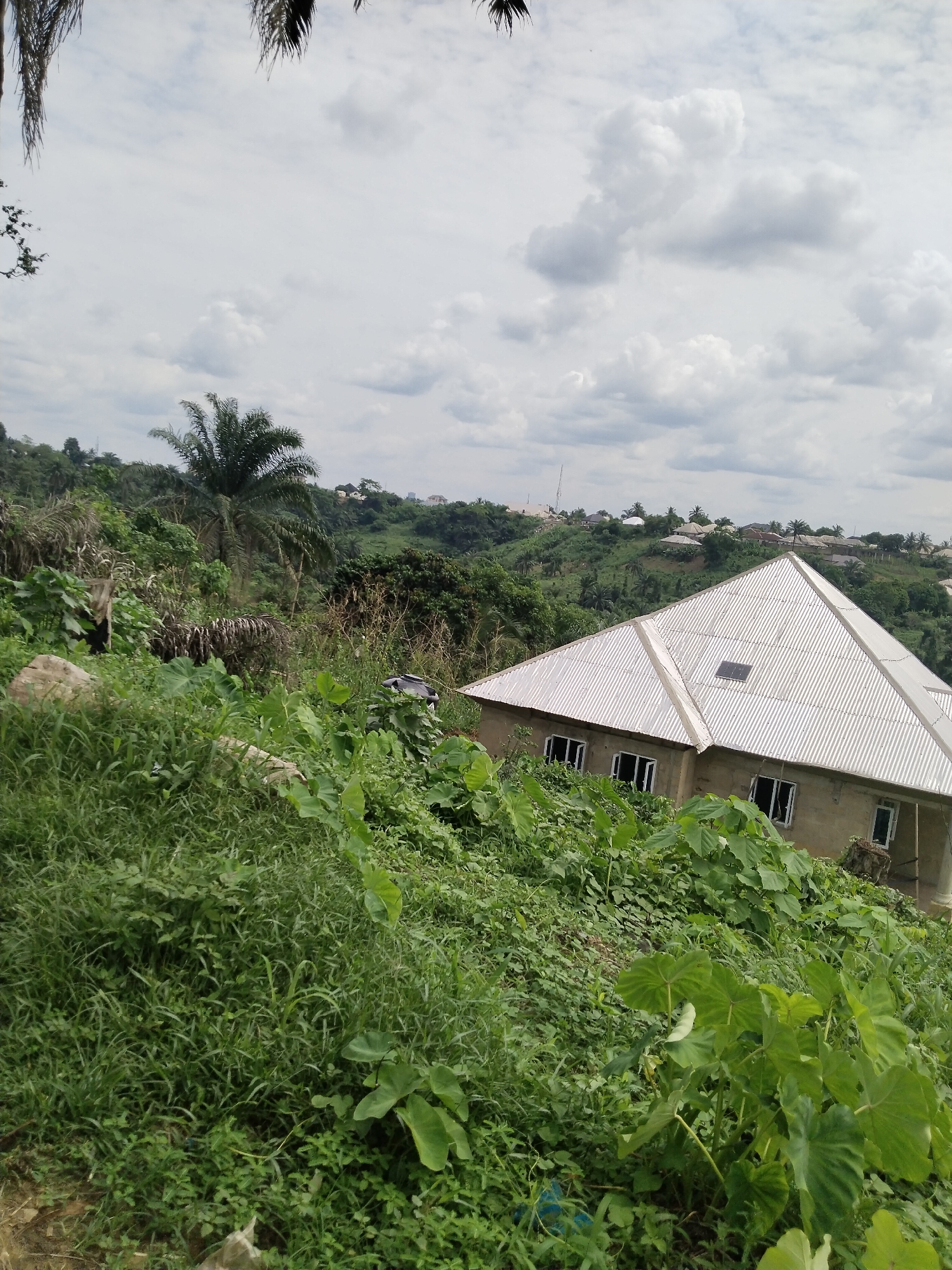 A view of a house under construction with a corrugated metal roof in Uyo, Nigeria. The house is partially obscured by overgrown vegetation. The background features a hilly landscape with scattered trees and buildings. The sky is cloudy. There are no people visible.