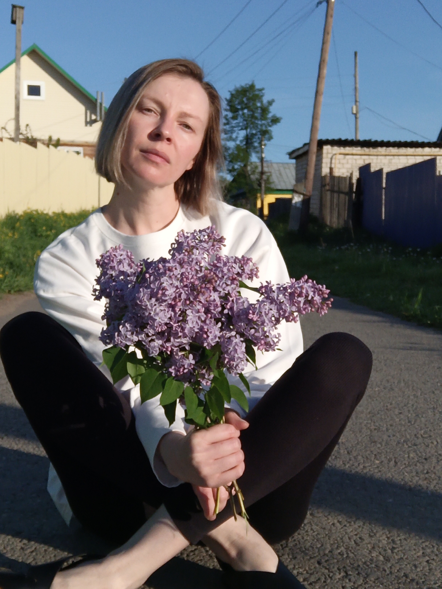 A medium shot depicts a fair-skinned woman with short, blonde hair, sitting cross-legged on an asphalt road. She is wearing a white, long-sleeved shirt and black leggings. In her hands, she holds a bouquet of purple lilacs. The woman appears to be looking directly at the viewer with a neutral expression.
The background reveals a rural street scene with various buildings. To the left, there is a light yellow house with a green roof and a window. A yellow fence runs along the edge of the property. To the right, there is another building with a dark blue fence and utility poles can be seen in the distance. Green grass lines the sides of the road.
The location is speculated to be in the city of Chaikovsky, Russia.