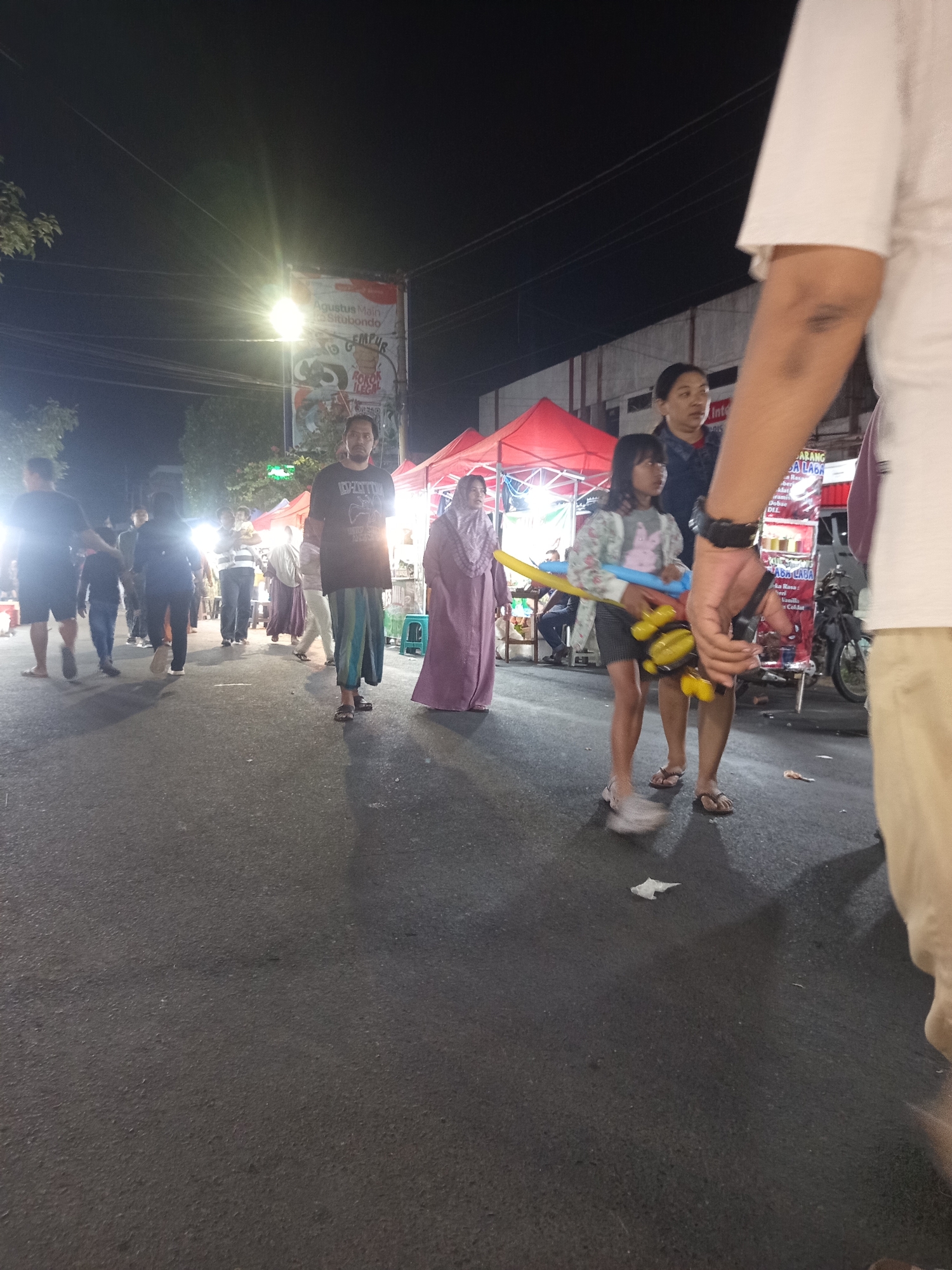 The image depicts a night scene in Ardirejo, Indonesia, likely a street market or festival. Several people are walking along an asphalt road. In the foreground, a person wearing a white shirt and beige pants is partially visible on the right. A young girl in the middleground is holding a yellow and blue balloon animal, and a woman in a purple headscarf stands to her left. Behind them, several other individuals are walking, some in groups. Red tents are set up on the right side of the road, suggesting stalls or vendor booths. A large poster with Indonesian text is visible in the background, illuminated by a bright streetlamp. The overall atmosphere appears lively and busy.