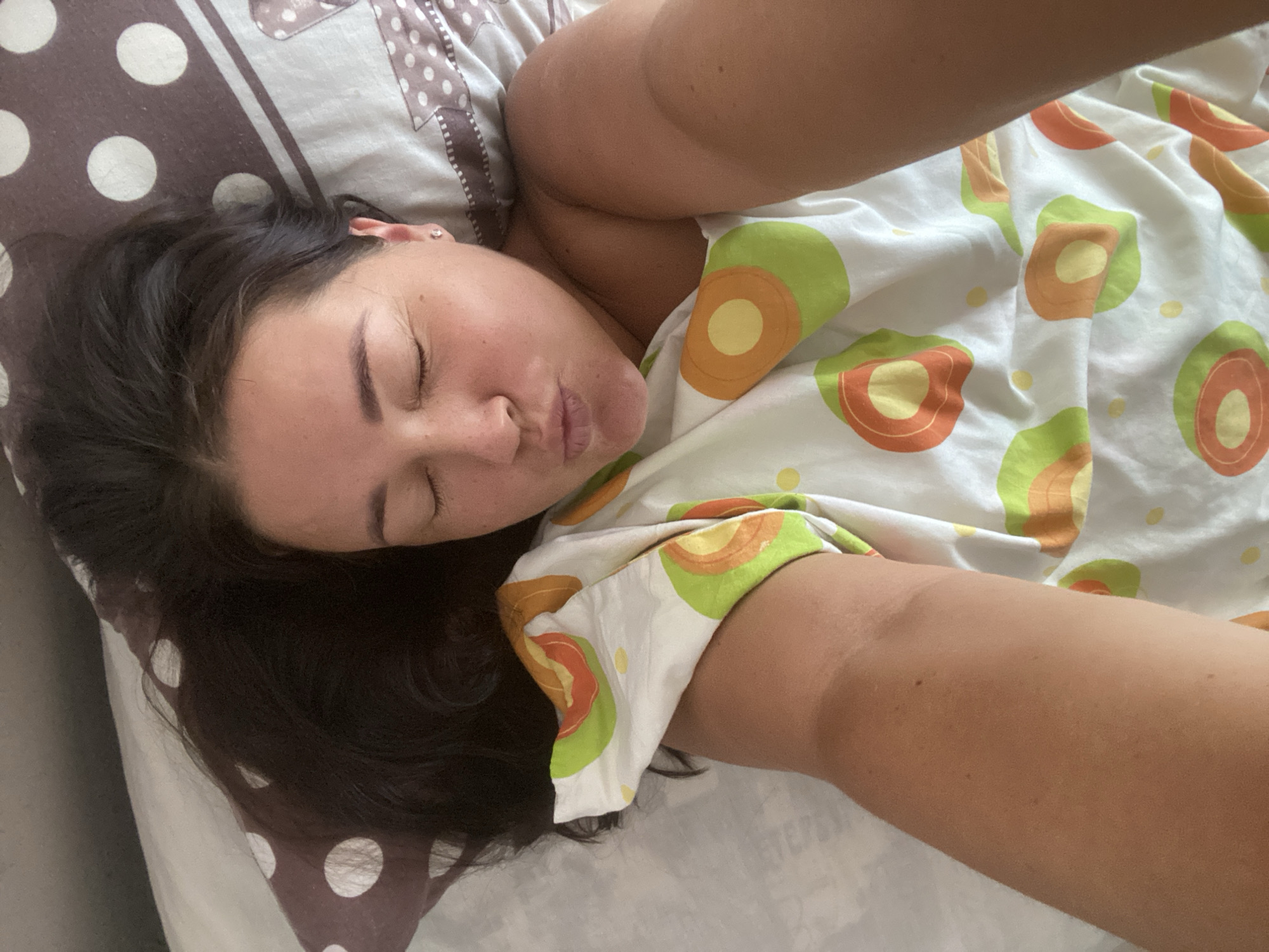 A woman with dark hair and closed eyes is lying in bed, with her face turned towards the camera in a pout. She is covered by a white blanket with a pattern of green and orange avocado slices. The pillow she is resting her head on has a brown and white polka dot pattern. The scene appears to be indoors, in a bedroom. The image was taken in Sochi, Russia.
