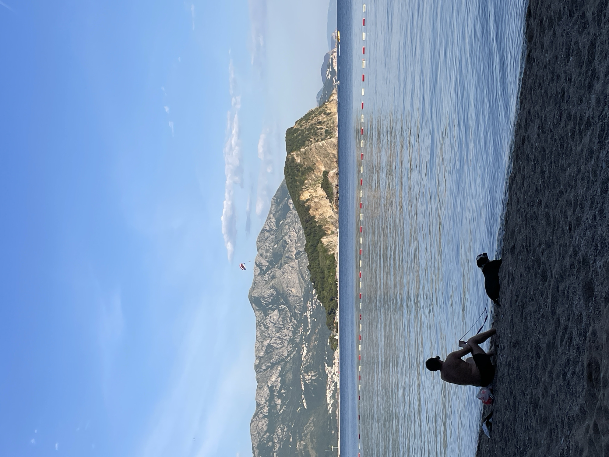 A horizontal outdoor shot depicts a beach scene in Budva, Montenegro. The sky is blue with some clouds, and the sea is calm. A mountain range is visible in the background with a parasailor in the sky. A line of red and white buoys is positioned in the sea. On the beach, a man sits while holding a leash connected to a black dog.