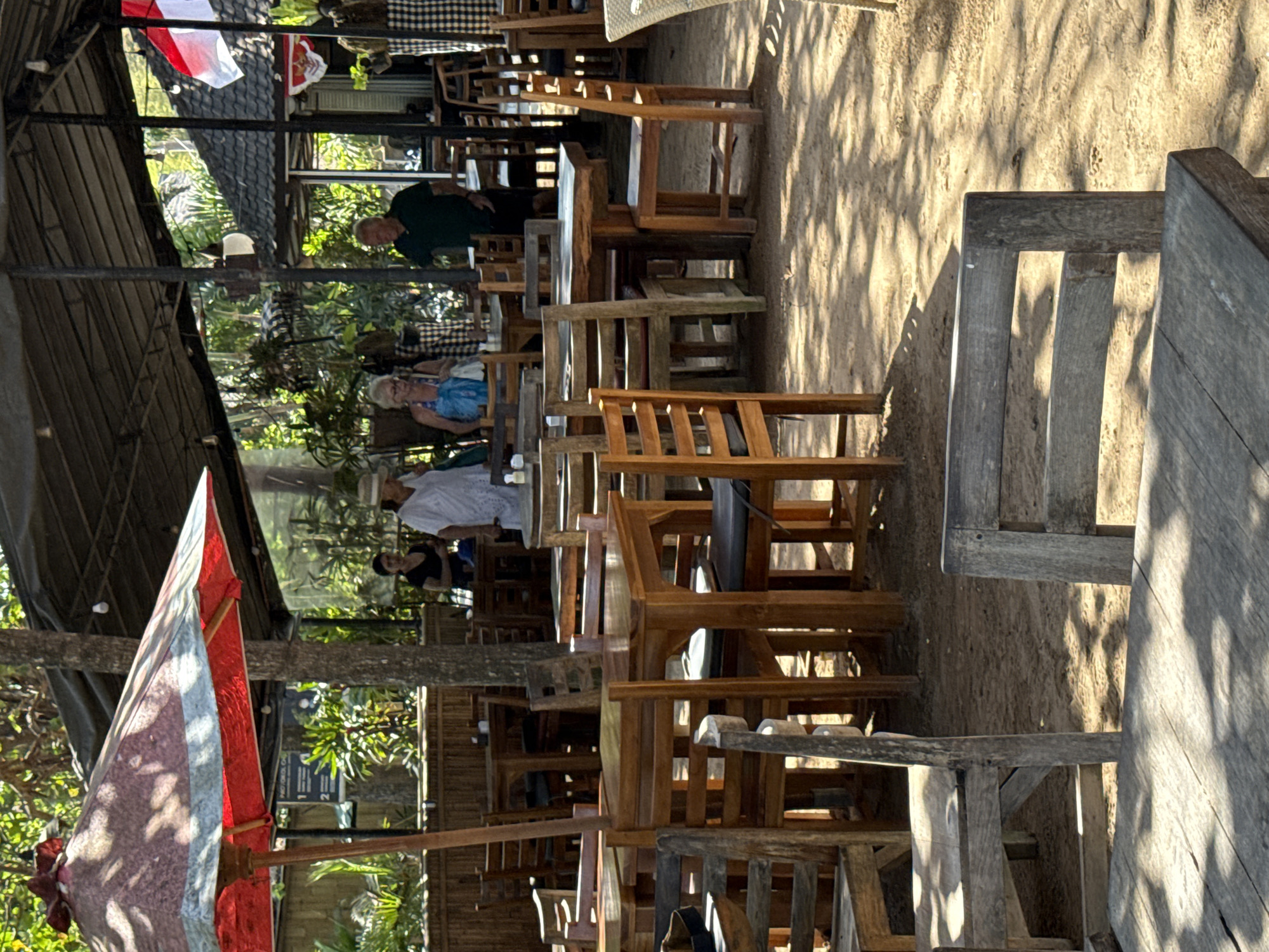 This is a medium-shot photograph of an outdoor seating area of a restaurant or cafe. The scene is set in Denpasar, Indonesia, as indicated by the Indonesian flags present. The environment is sandy, with dappled sunlight filtering through trees, creating shadows on the ground and tables.
In the foreground, several wooden tables and chairs are arranged. The furniture appears weathered and rustic. Some chairs have dark cushions. A large, red and white striped umbrella with a wooden pole stands to the left, providing shade over a table.
In the midground, several people are visible. A woman in a white dress and a straw hat is standing on the left. To her right, a woman wearing a blue swimsuit cover-up and sunglasses is seated. Further to the right, a man in a dark green polo shirt and shorts is standing. They appear to be patrons of the establishment, possibly waiting for or having finished their meals. Behind them, more tables and chairs are visible, as is the structure of the cafe, including a dark-colored roof supported by poles.
In the background, there are more trees and vegetation, suggesting a tropical setting. Two Indonesian flags, a large one and a smaller one, are displayed on the right side of the image. The overall atmosphere is casual and relaxed, indicative of a tropical dining experience.